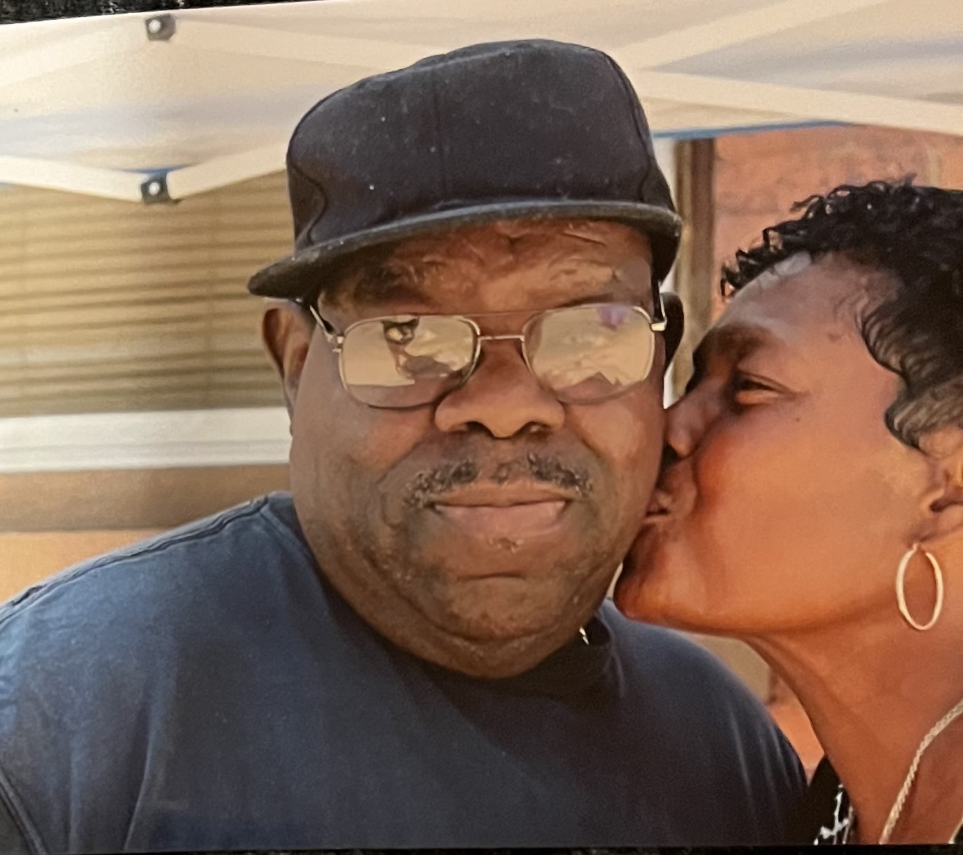 Person wearing glasses and a black cap receives a kiss on the cheek from a woman with short curly hair and hoop earrings.