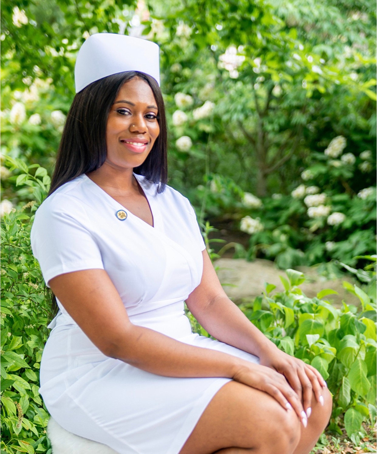 Woman in white nurse uniform and hat sitting outdoors with green foliage background.