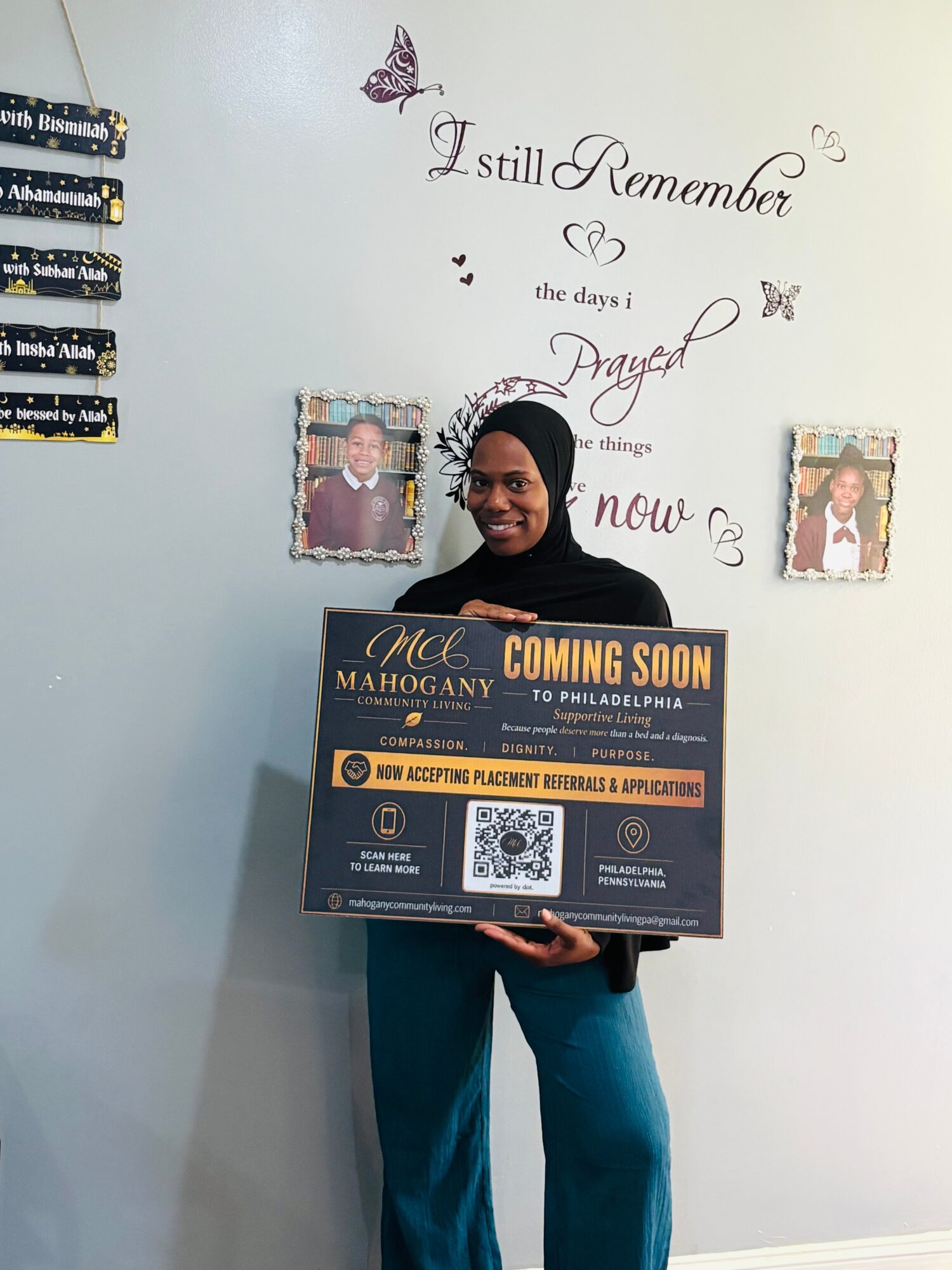 Person holding a sign in front of a wall with decorative text and butterflies, smiling, wearing a black headscarf and blue pants.