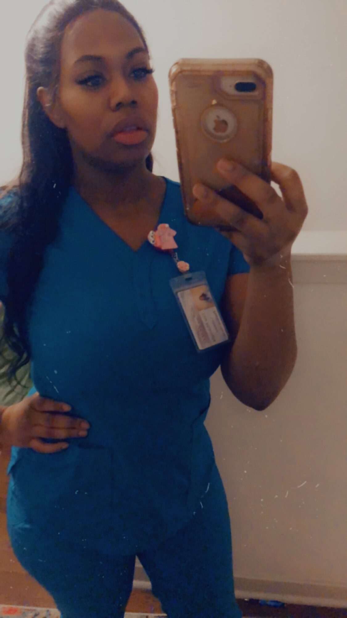 Person in blue scrubs taking a mirror selfie with a phone, hand on hip, neutral background.