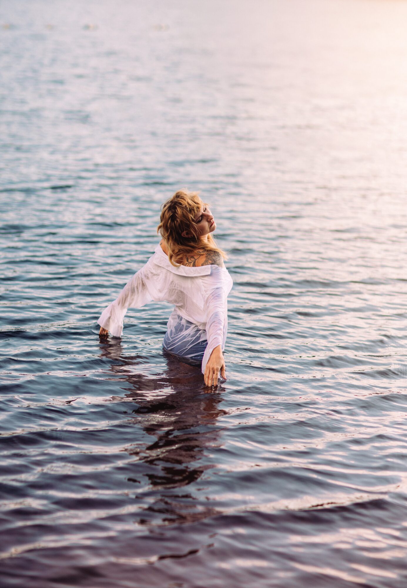 Person standing in water, facing away, with hair and white shirt, water reaching mid-thigh, calm water surface.