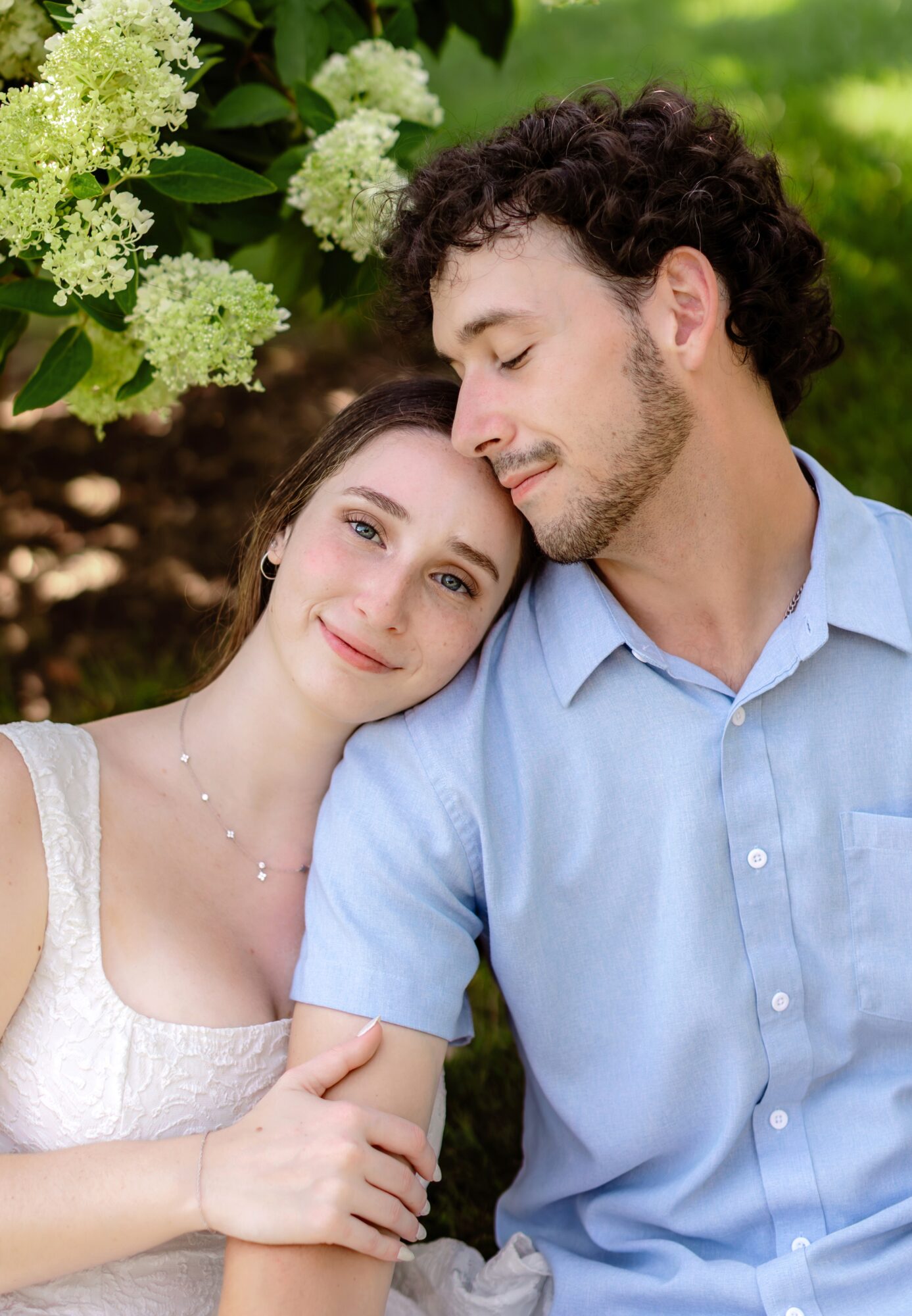 A couple outdoors, smiling and leaning on each other, surrounded by greenery and white flowers.