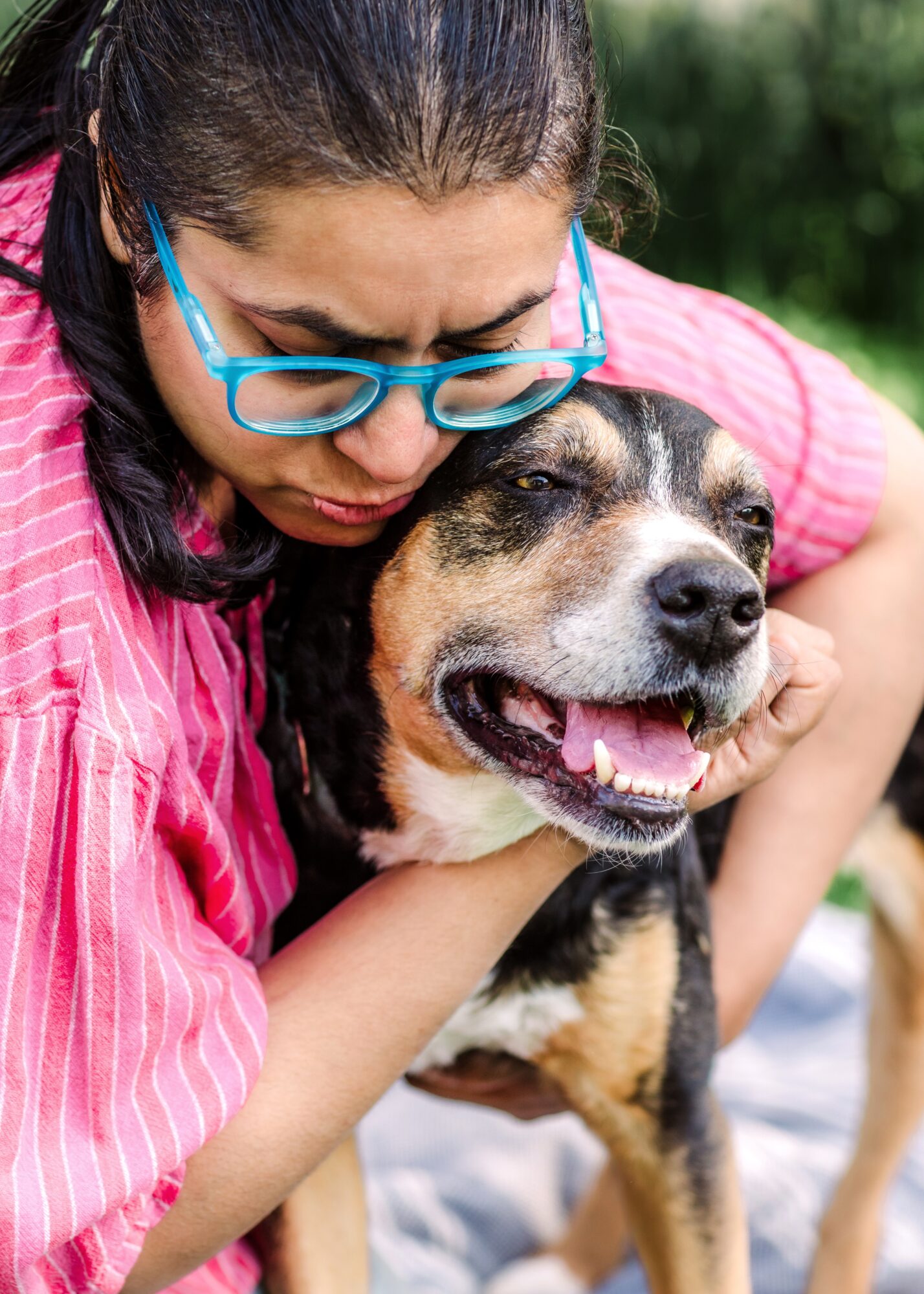 Person hugging a happy dog outdoors, person wearing glasses and pink shirt, dog with closed eyes and open mouth.