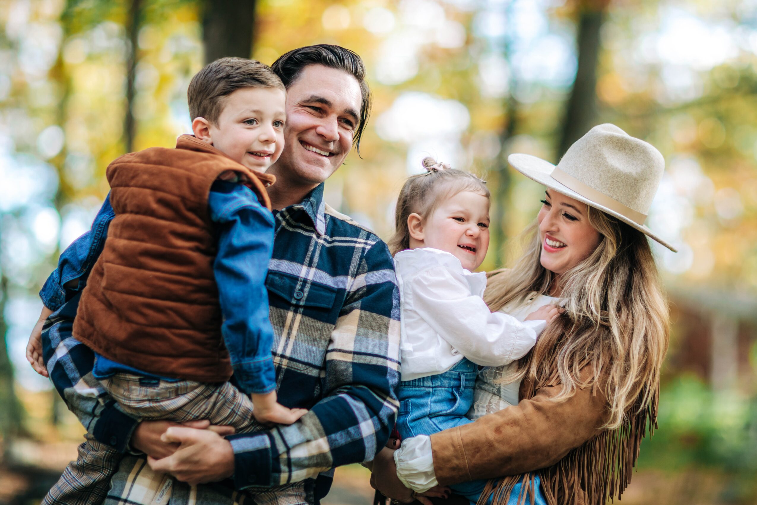 Family of four outdoors, smiling, with trees and sunlight in background.