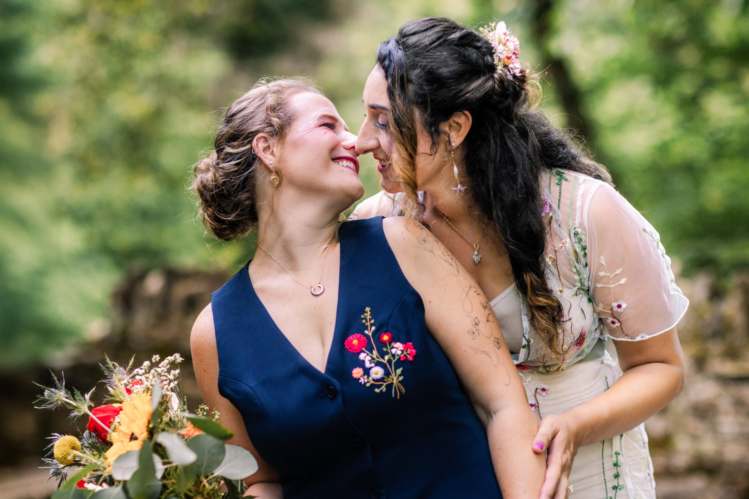 Two women smiling and touching noses outdoors, one holding a bouquet, surrounded by green trees.