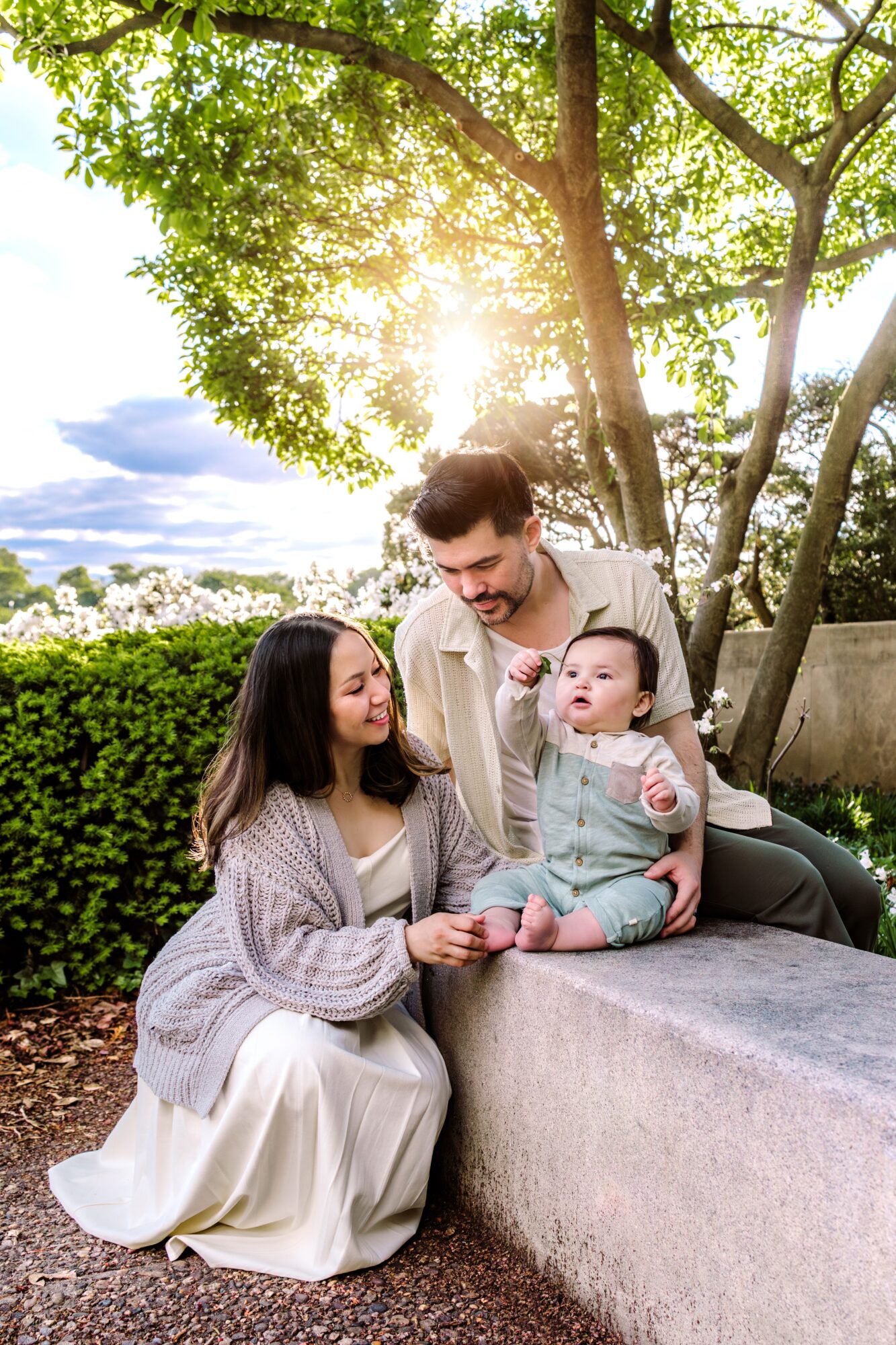 Family of three outdoors, woman sitting, man standing, baby on a stone ledge, sunlight through trees, green foliage, cloudy sky