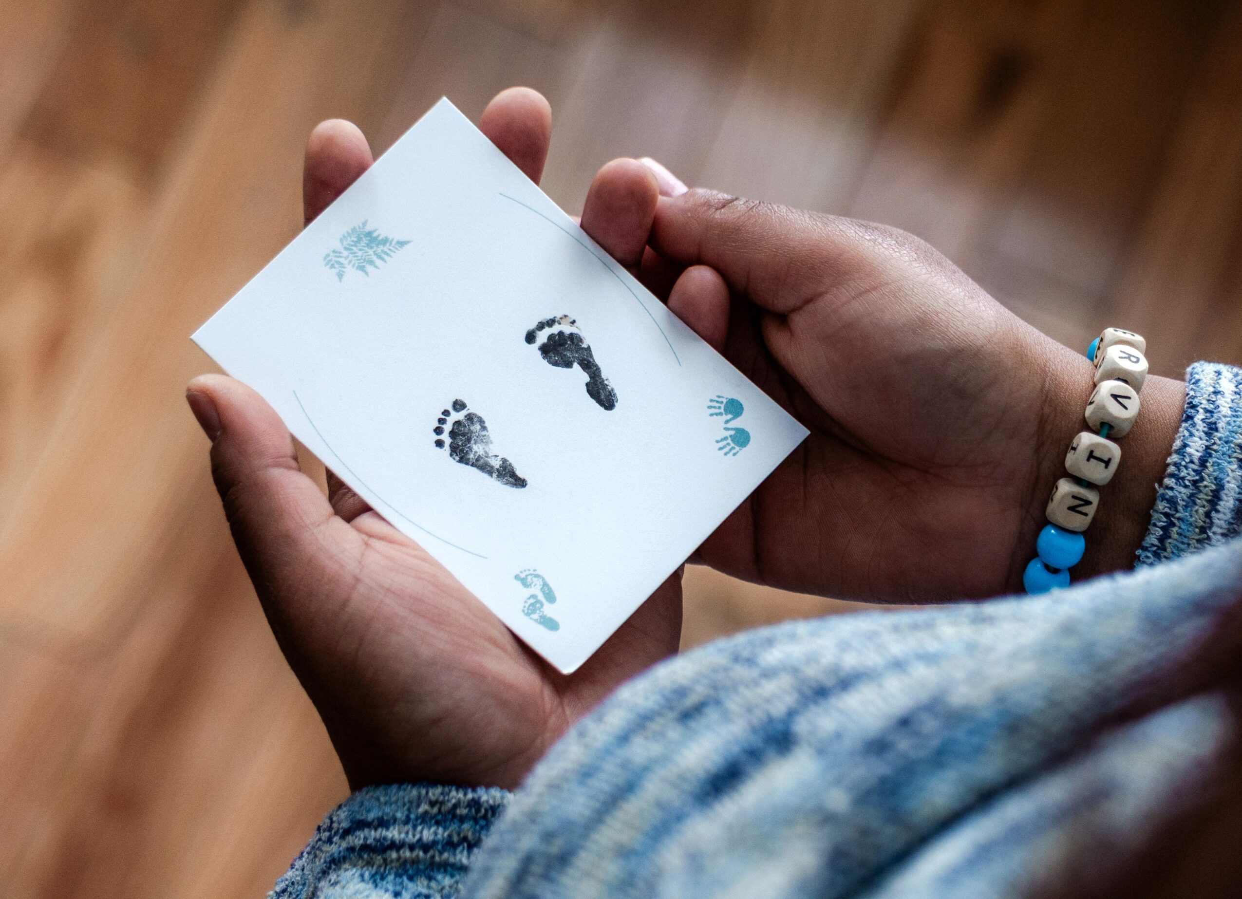 Person holding a card with baby footprints and handprints, wearing beaded bracelets, with a blurred wooden background.
