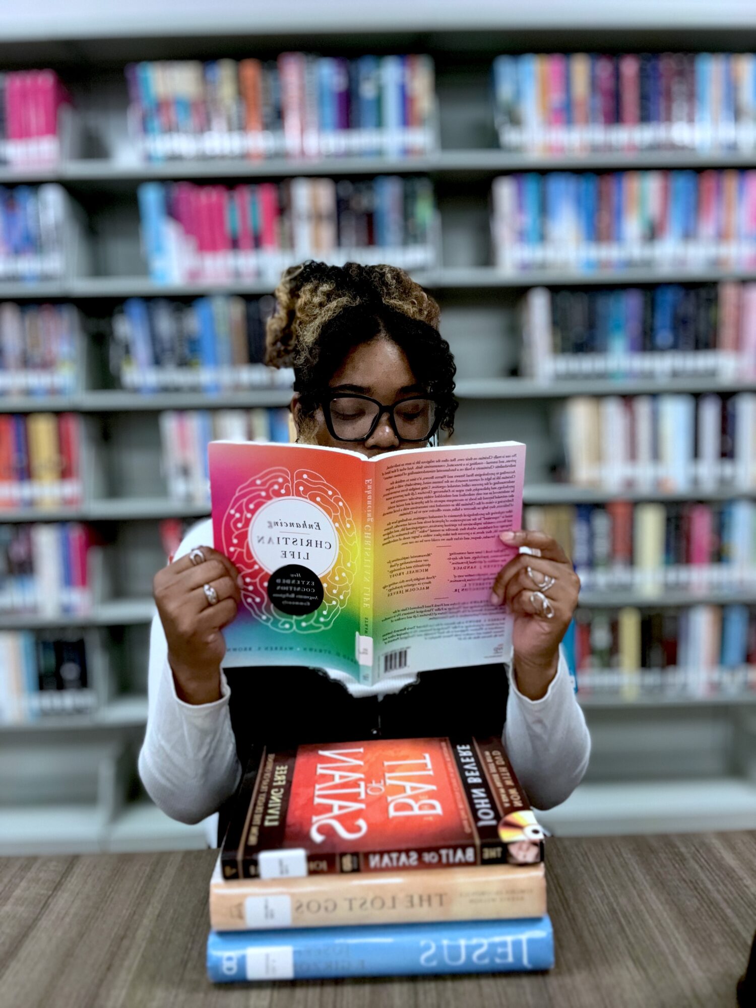 Person reading a book at a library table with shelves of books in the background.