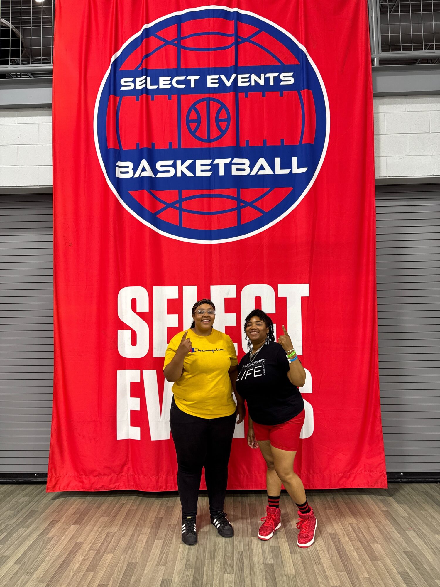 Two women stand in front of a large red banner with a basketball logo and text, smiling and posing indoors.