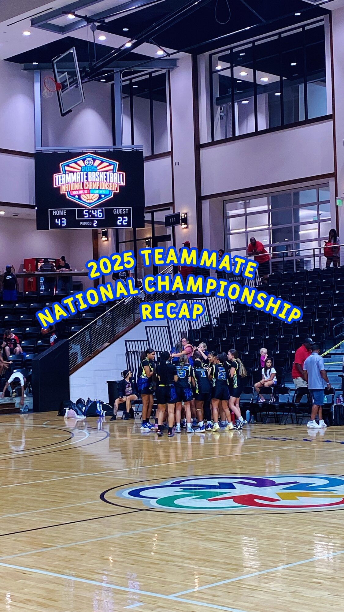 Basketball team huddled on court with scoreboard and banners in background, colorful logo on floor, indoor gym setting.
