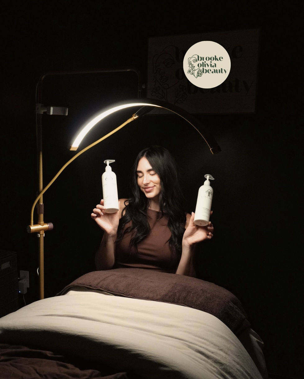 Woman holding two bottles, smiling, with a lamp and a screen in the background, dark setting, bed in foreground.