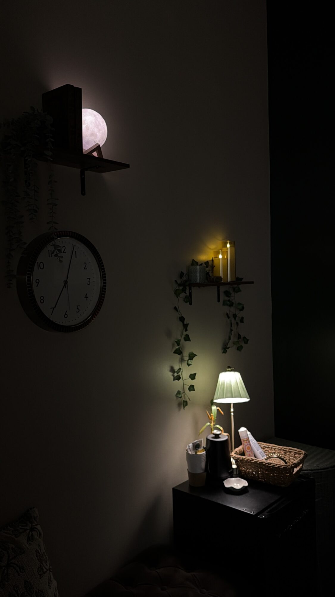 Dimly lit room with a wall clock, shelves with decorative items, and a table with a lamp and basket.