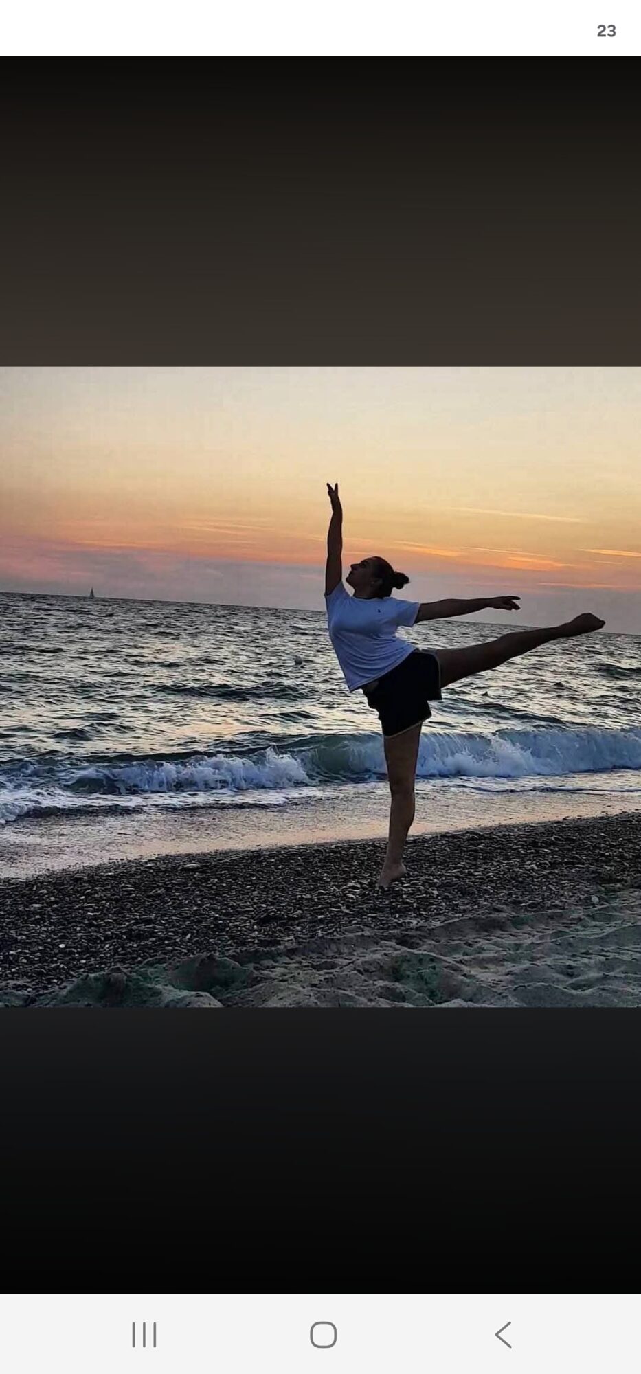 Person performing a ballet pose on the beach at sunset, with arms and leg extended, ocean waves in background.