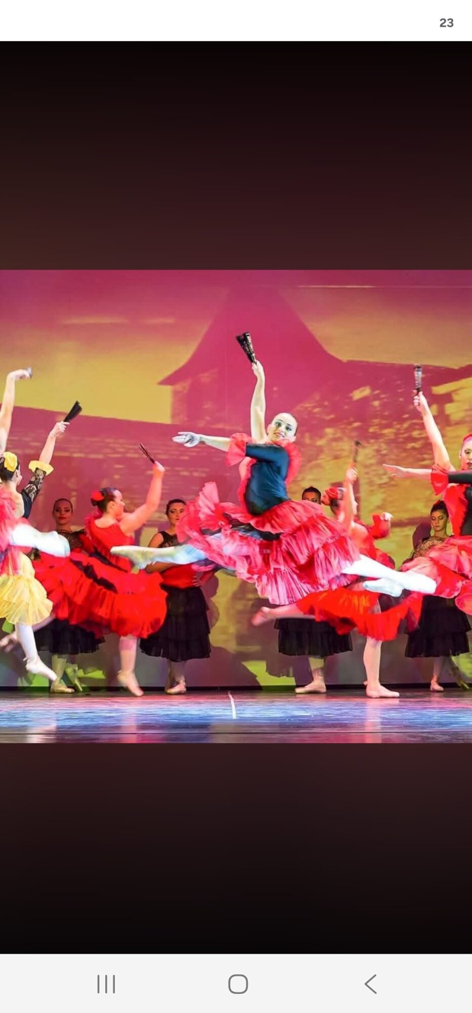 Group of dancers performing on stage with colorful costumes and a scenic backdrop, some raising arms, in a lively dance scene.