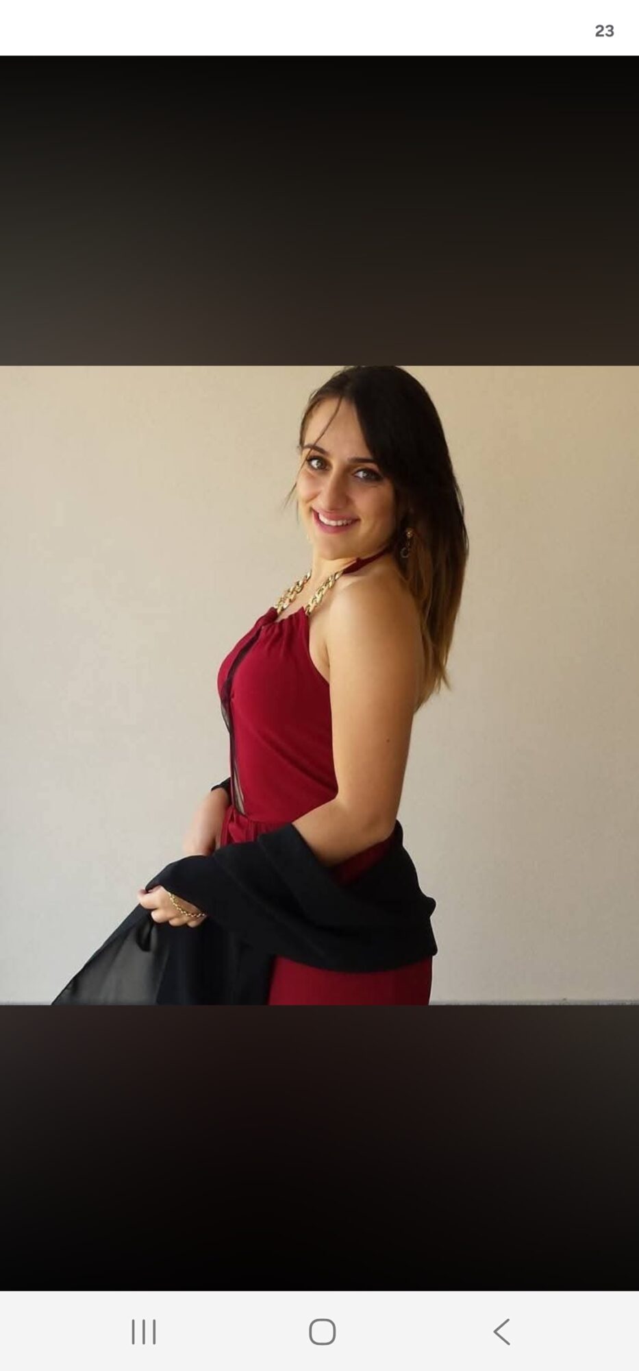 Woman with long dark hair wearing a red sleeveless top, smiling, holding a black jacket, standing against a plain background.