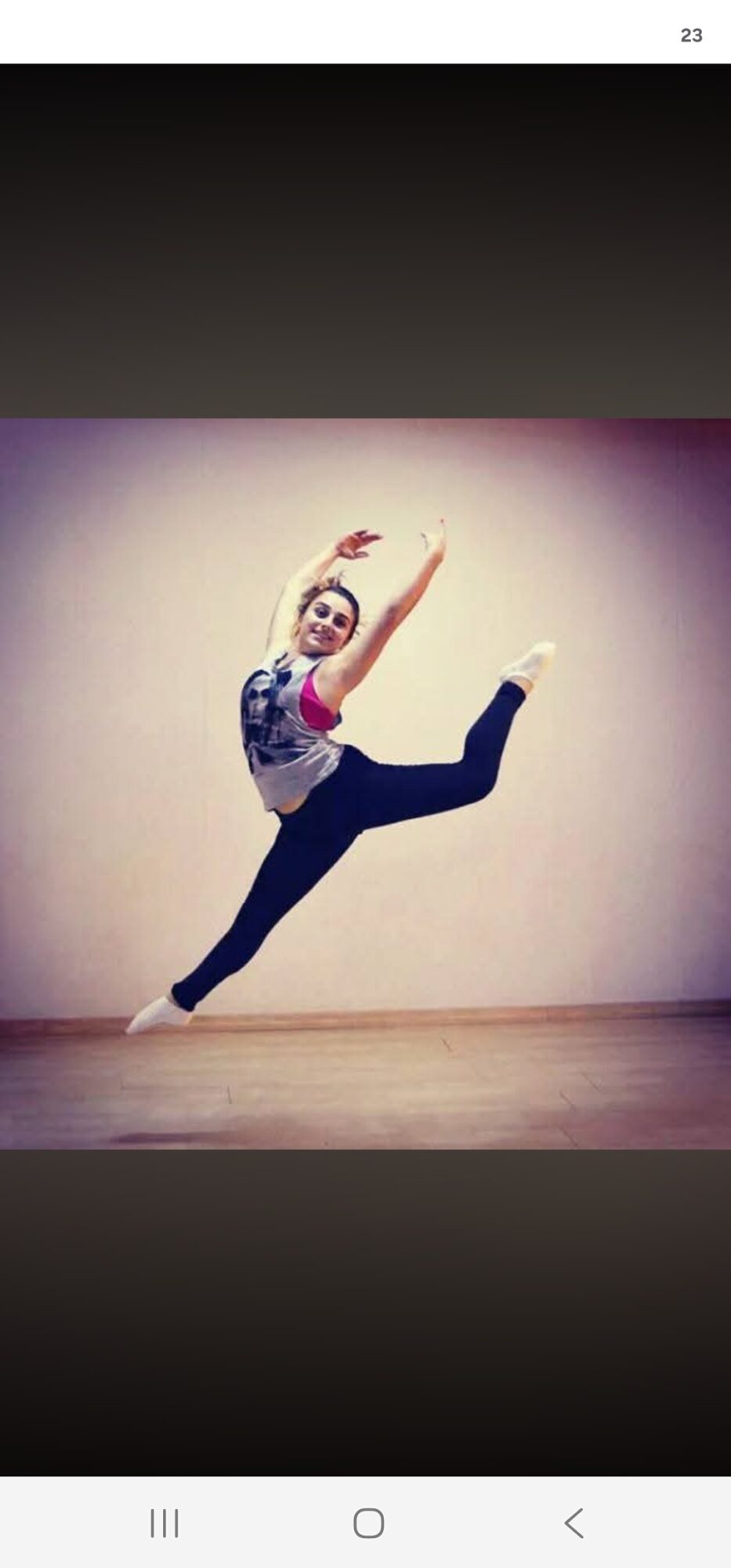 Person performing a dance move with one leg extended and arms raised, against a plain wall background.