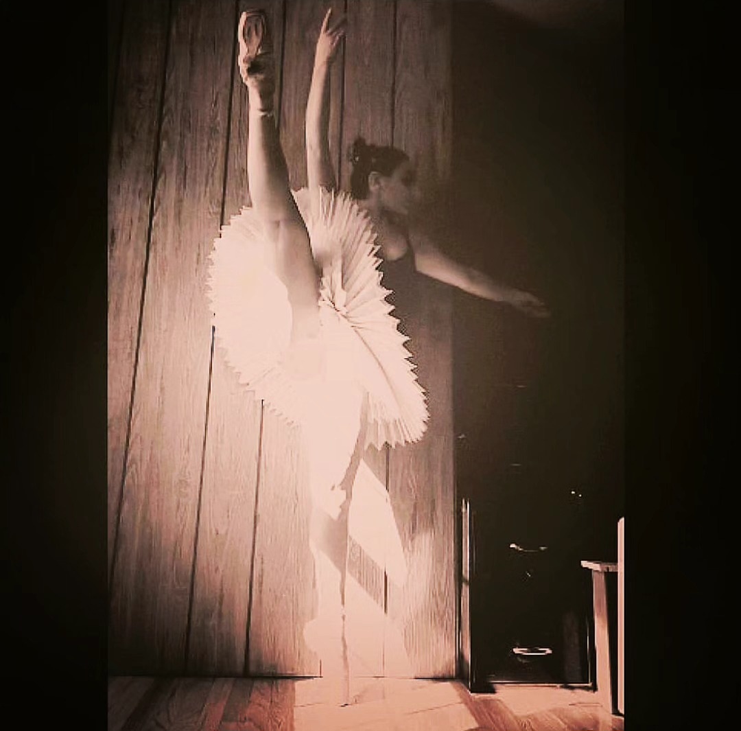 Ballet dancer in a tutu stretching with one leg on the wall, arms raised, on a wooden floor and wall.