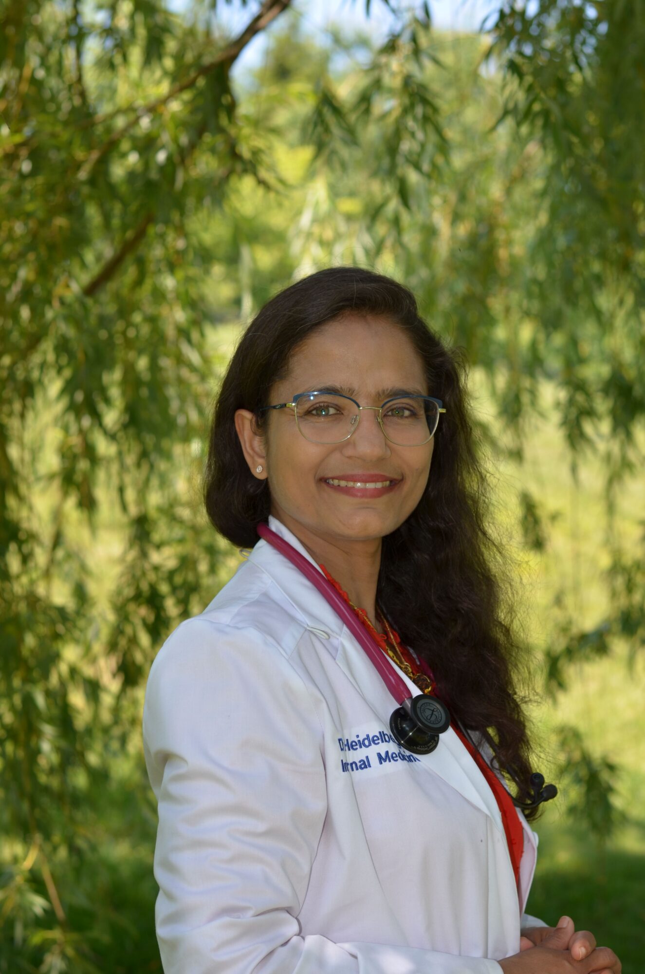 Female doctor outdoors with trees in background, wearing white coat and stethoscope, smiling at camera.