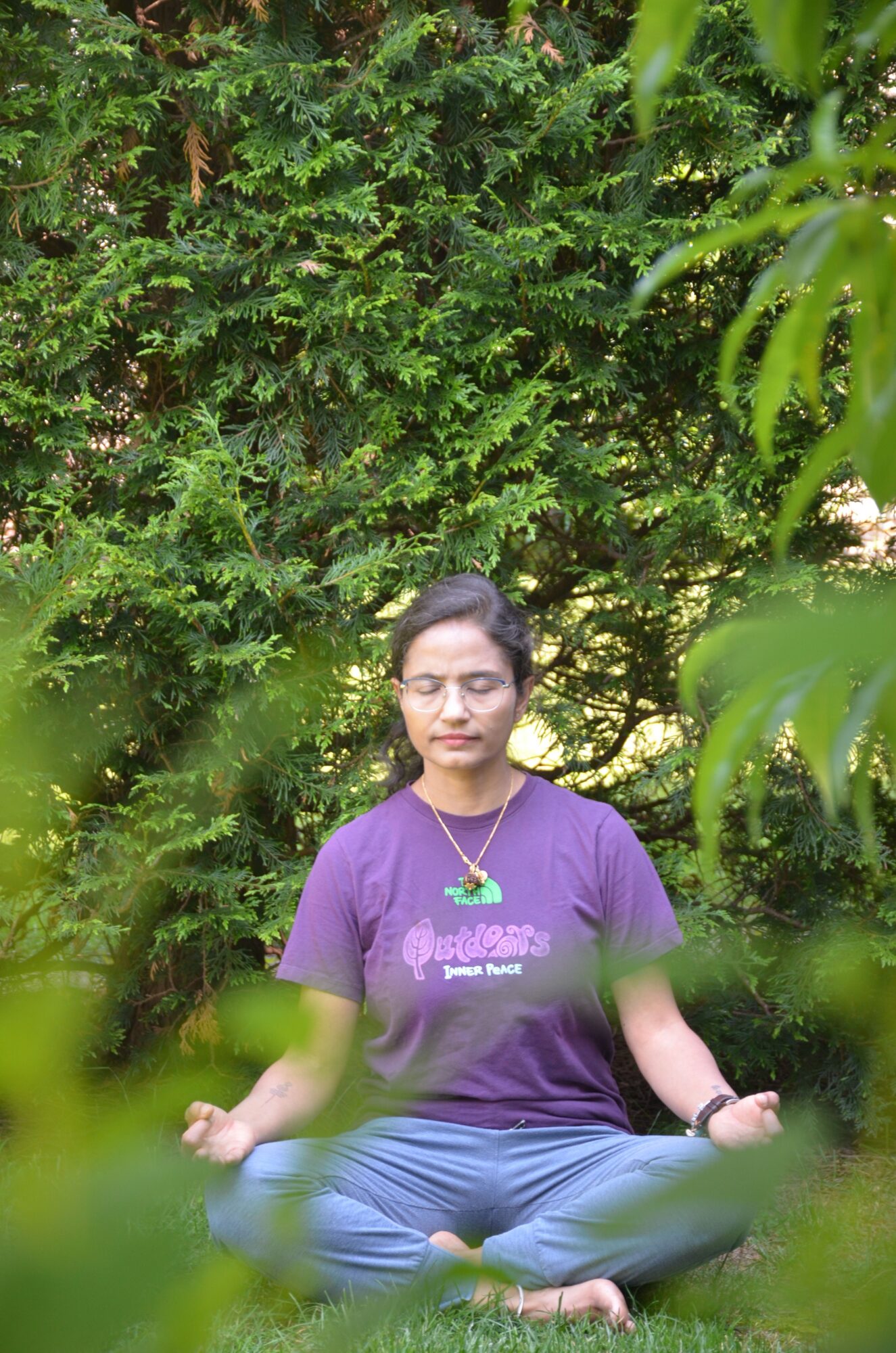 Woman sitting outdoors among green foliage, smiling, wearing a purple shirt and jeans.