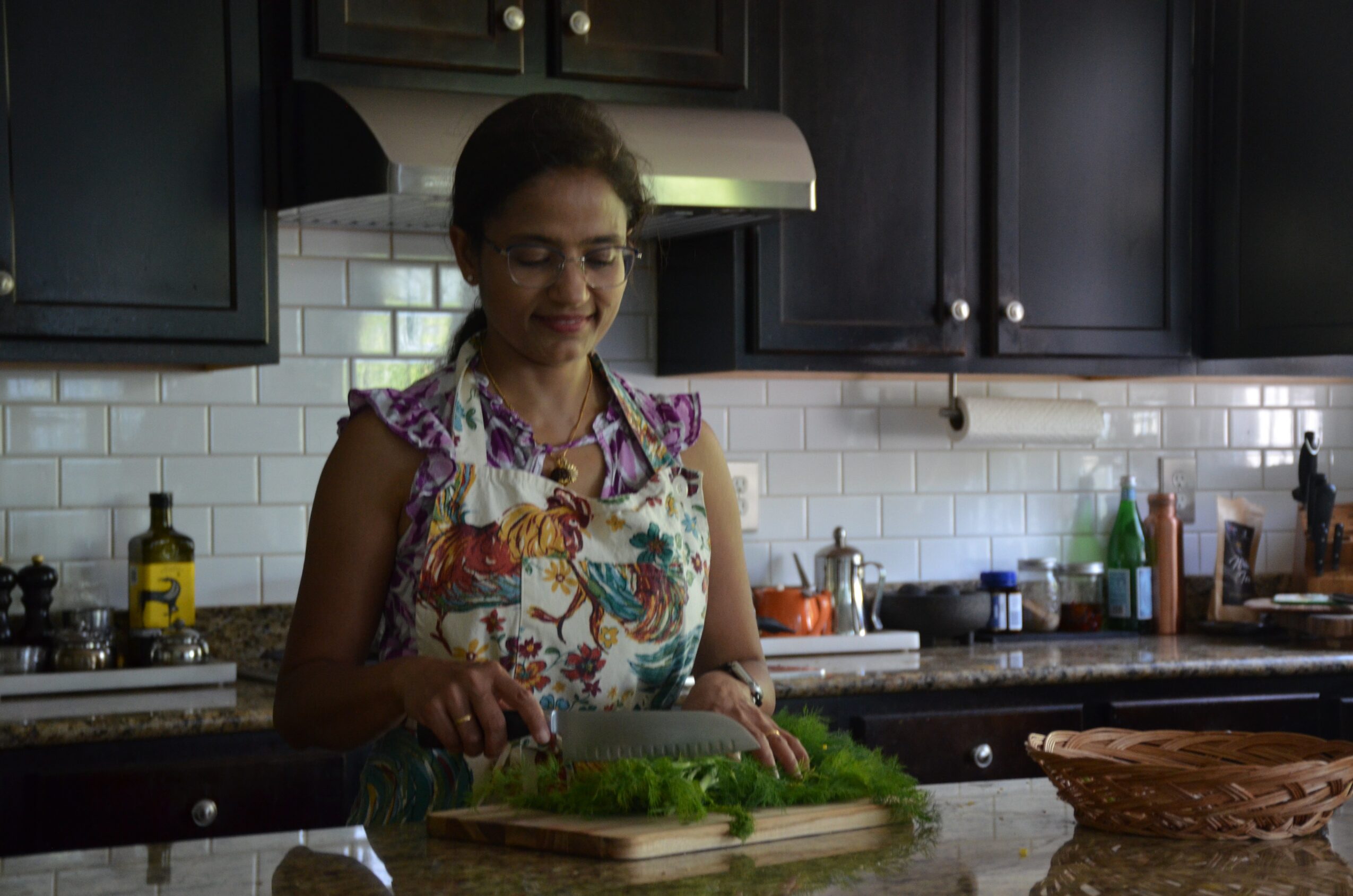 Woman in a floral apron preparing greens in a modern kitchen with dark cabinets and white subway tile backsplash.