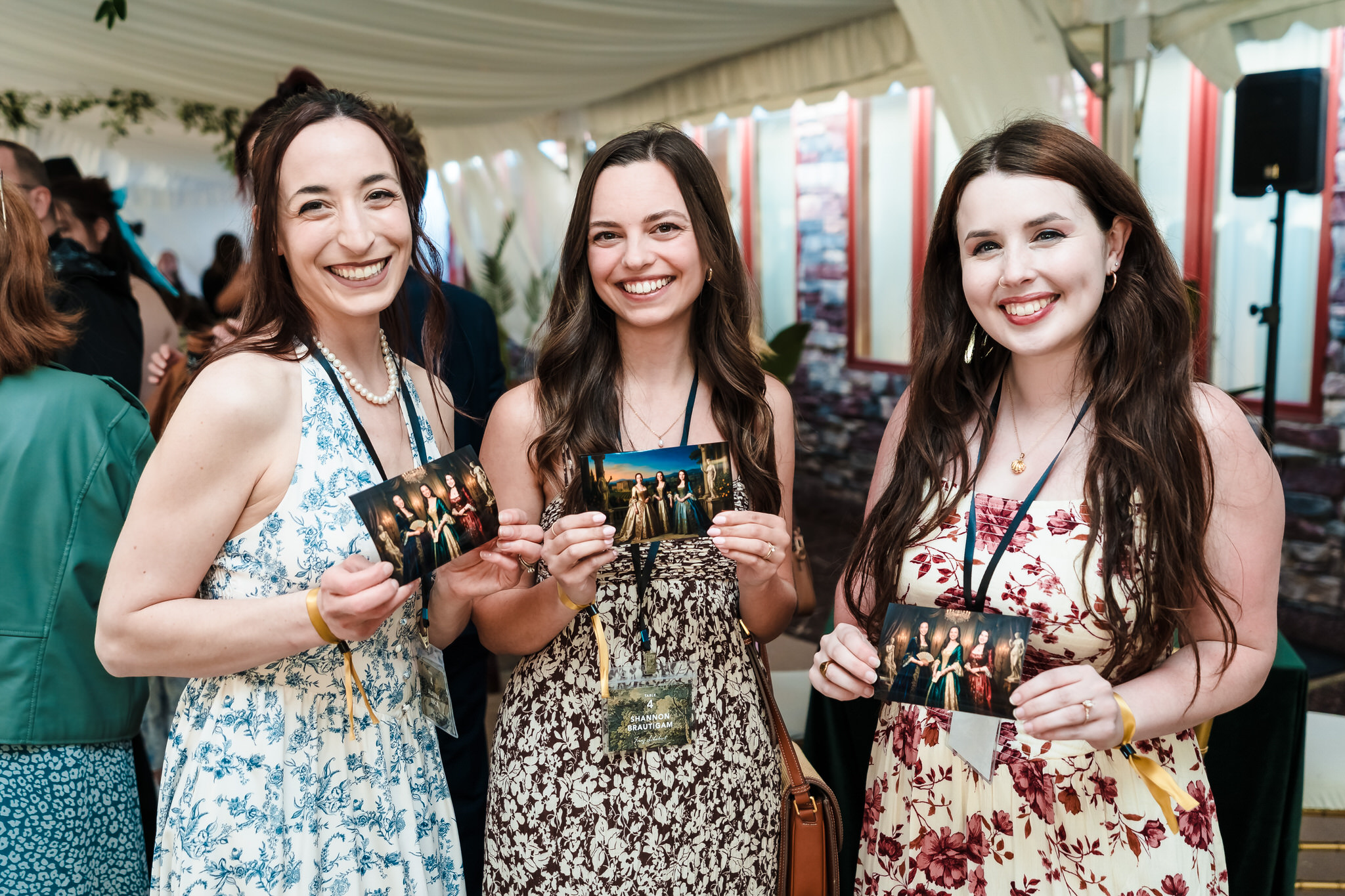 Three women smiling and holding photographs at an indoor event, with a tent and other people in the background.