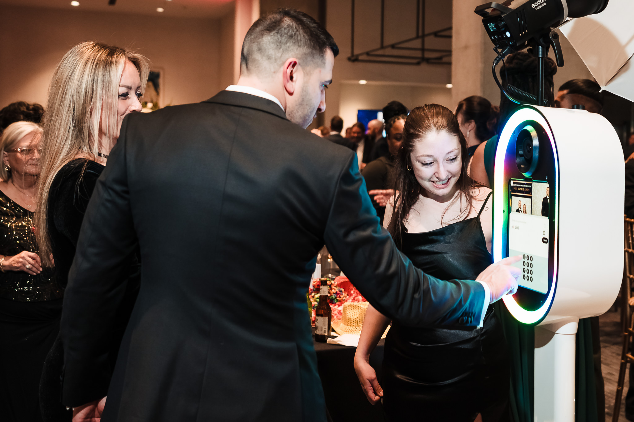 People gathered at an event, with a man in a suit using a photo booth, smiling girl nearby, and others in background.