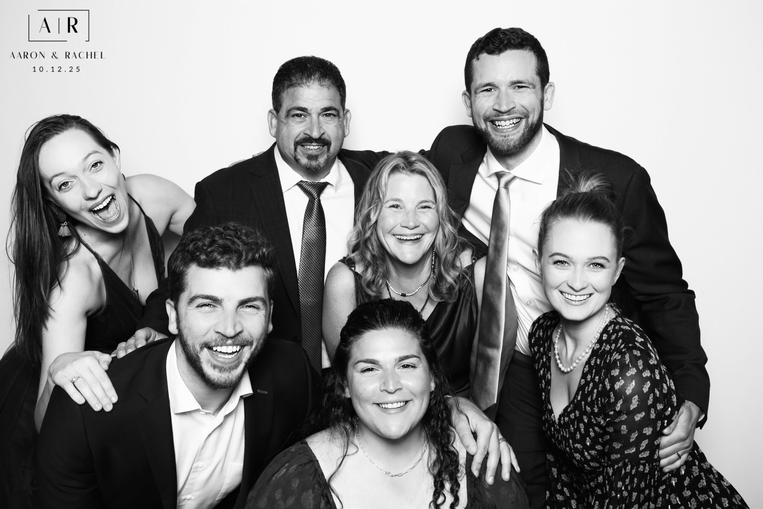 Group of seven people smiling and posing together in a photo, with some embracing and others standing close, in black and white.