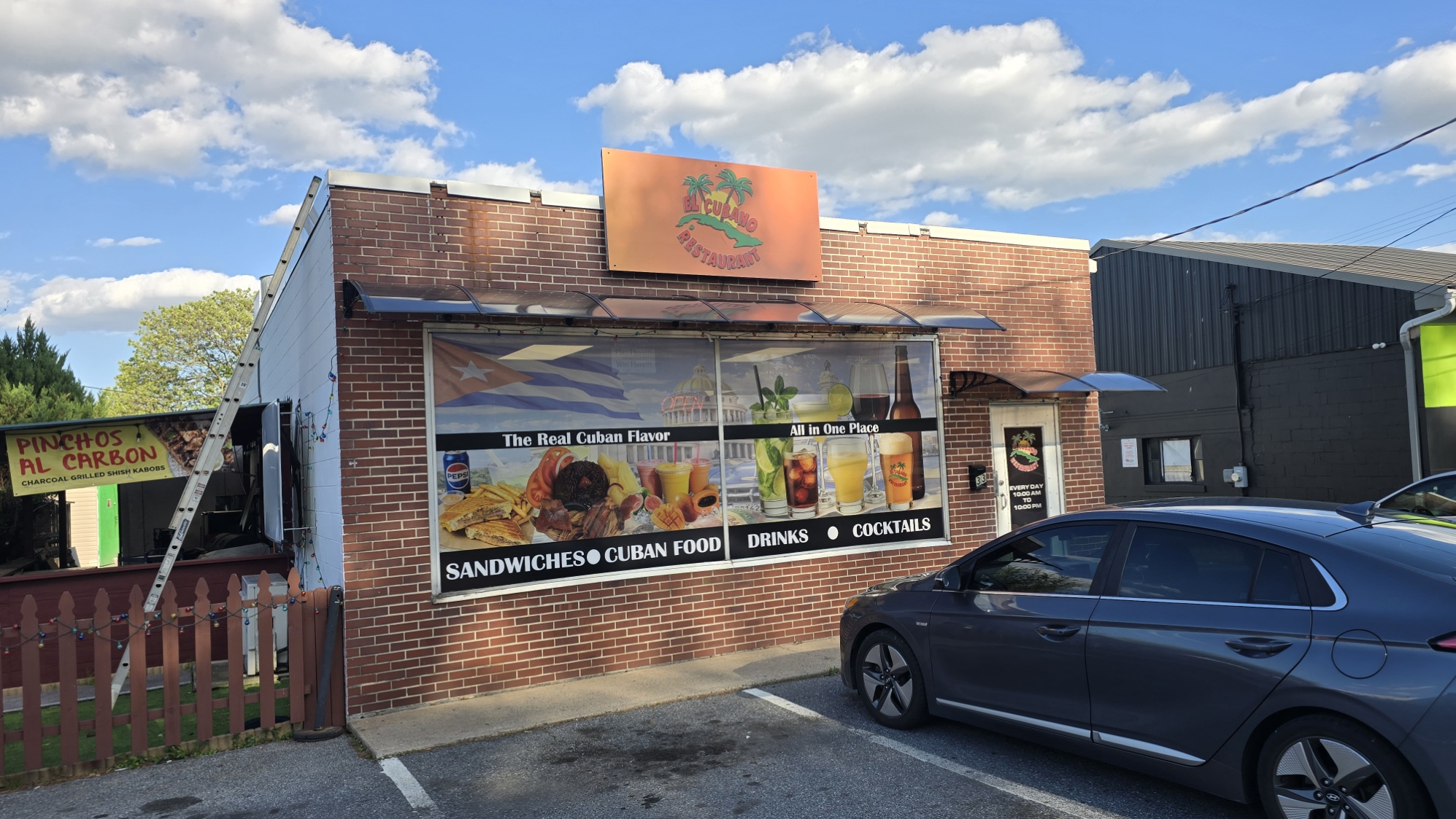 Brick building with a sign and window displaying food items, parked car in front, blue sky with clouds above.