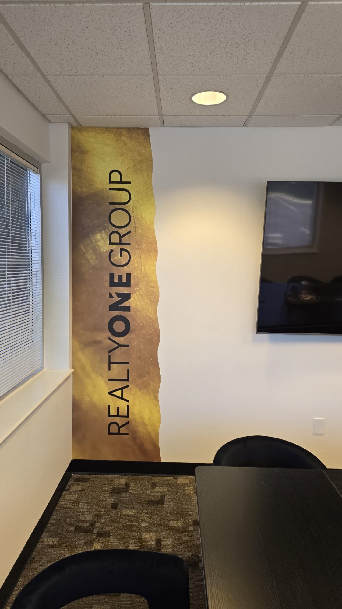 Interior view of a room with a wall-mounted TV, a window, and a banner reading 'REALTY ONE GROUP' on the wall.