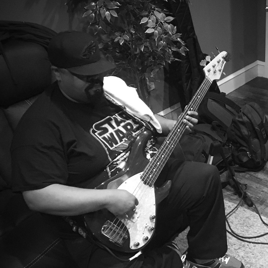 Person sitting on a couch playing an electric bass guitar, wearing a cap and glasses, with a plant and bags nearby.