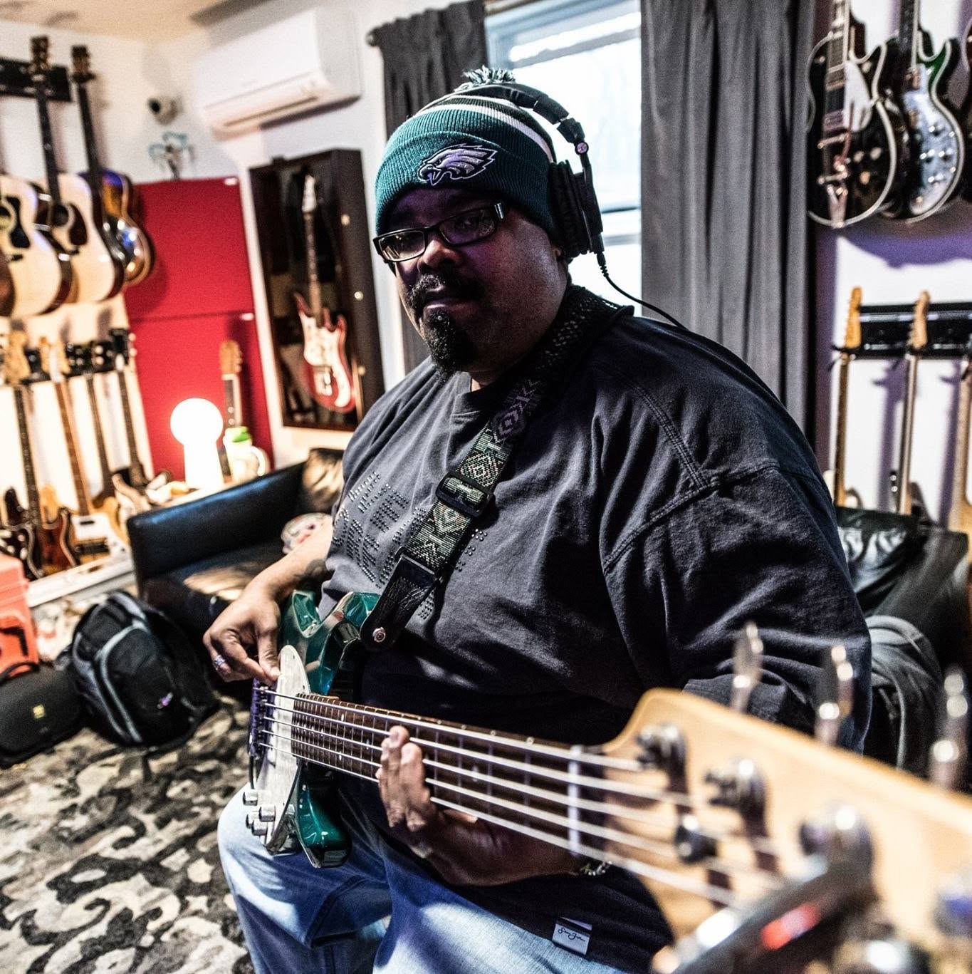 Person playing guitar in a music studio with guitars on the wall, wearing headphones and a beanie, surrounded by musical equipment.