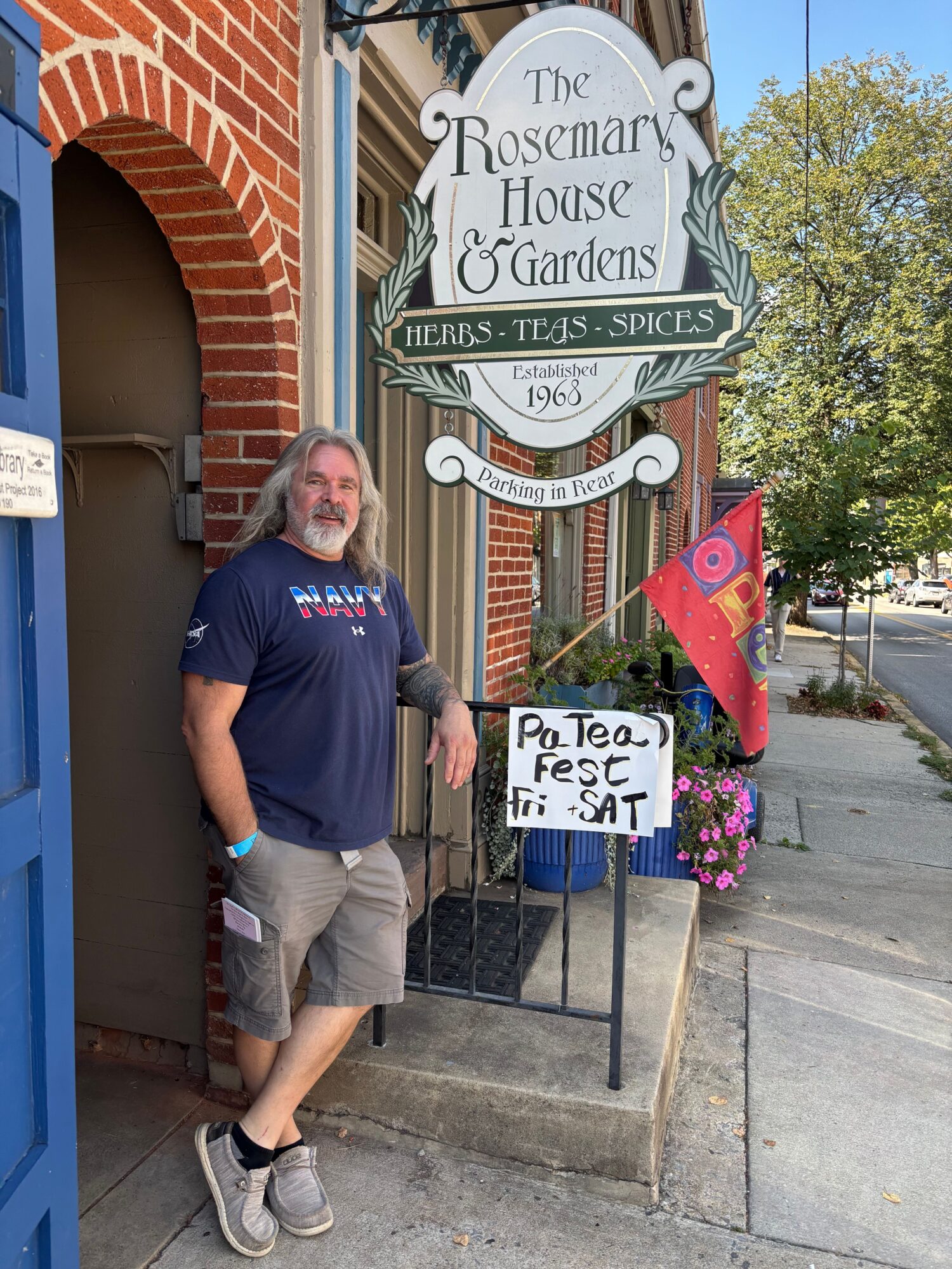 Man standing outside a building with a sign for The Rosamond H. Huffman Gardens, holding a sign for a team event, with flowers and sidewalk nearby.