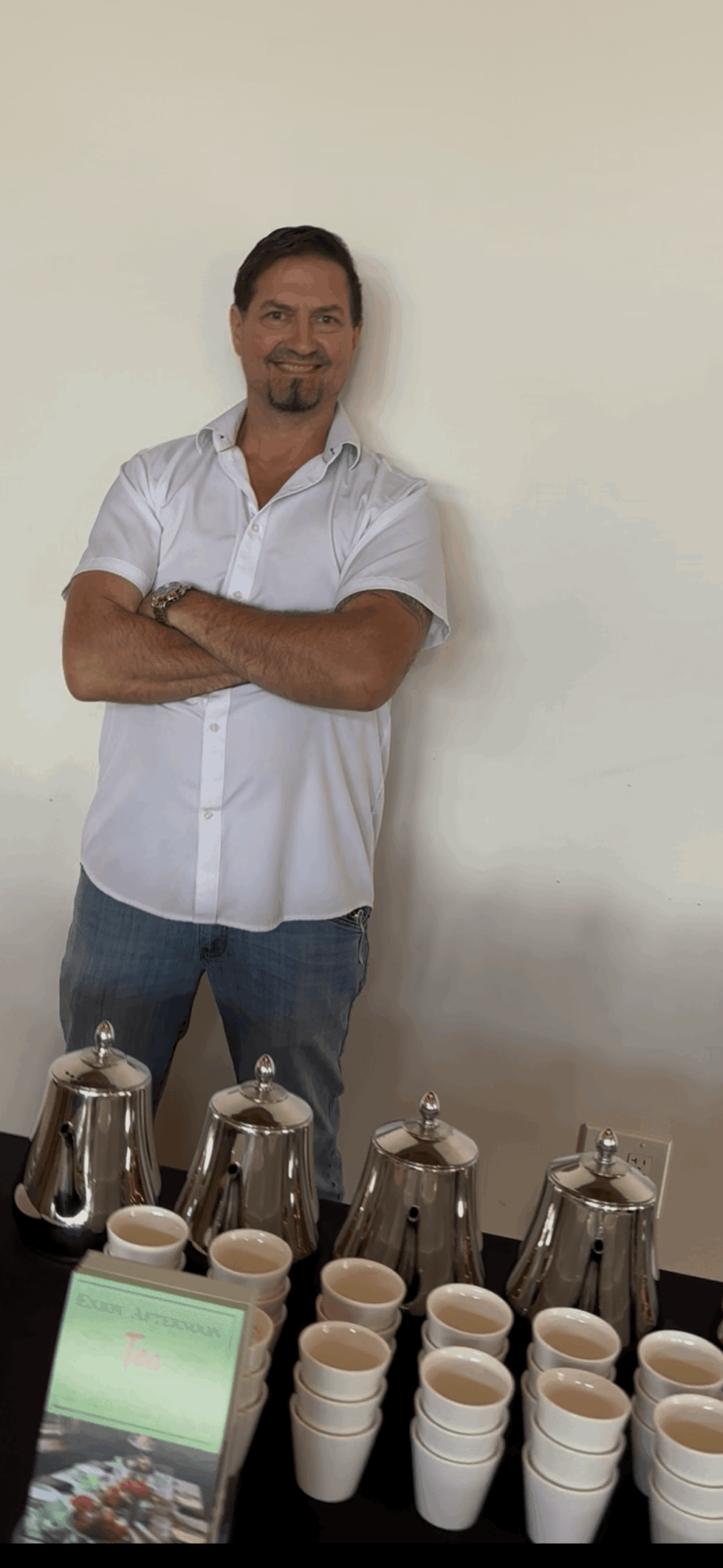 Man standing with arms crossed in front of a table with coffee cups and pots, smiling, against a plain wall.