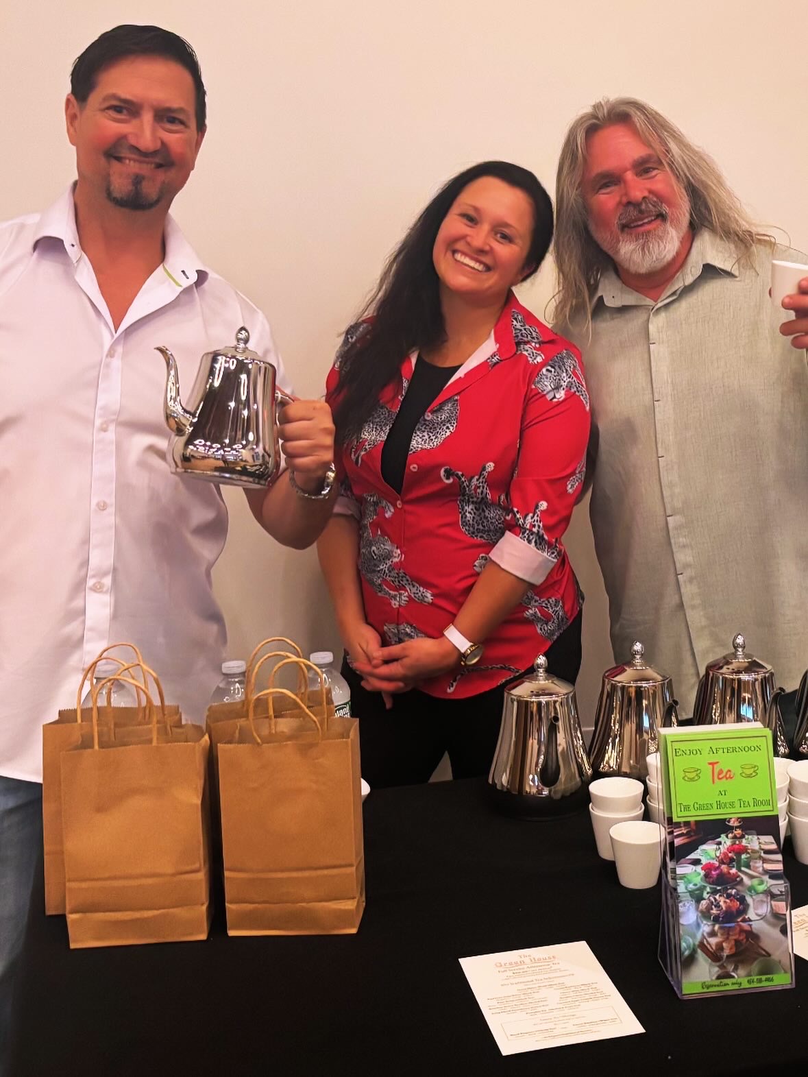 Three people smiling behind a table with gift bags, cups, and a brochure, indoors against a plain wall.