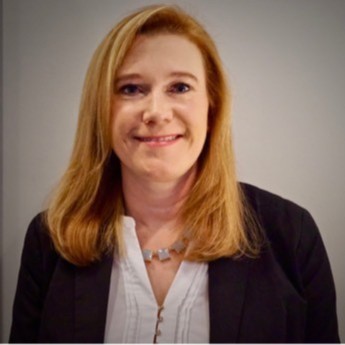 Portrait of a woman with shoulder-length blonde hair, smiling, wearing a black blazer and white blouse, against a plain background.