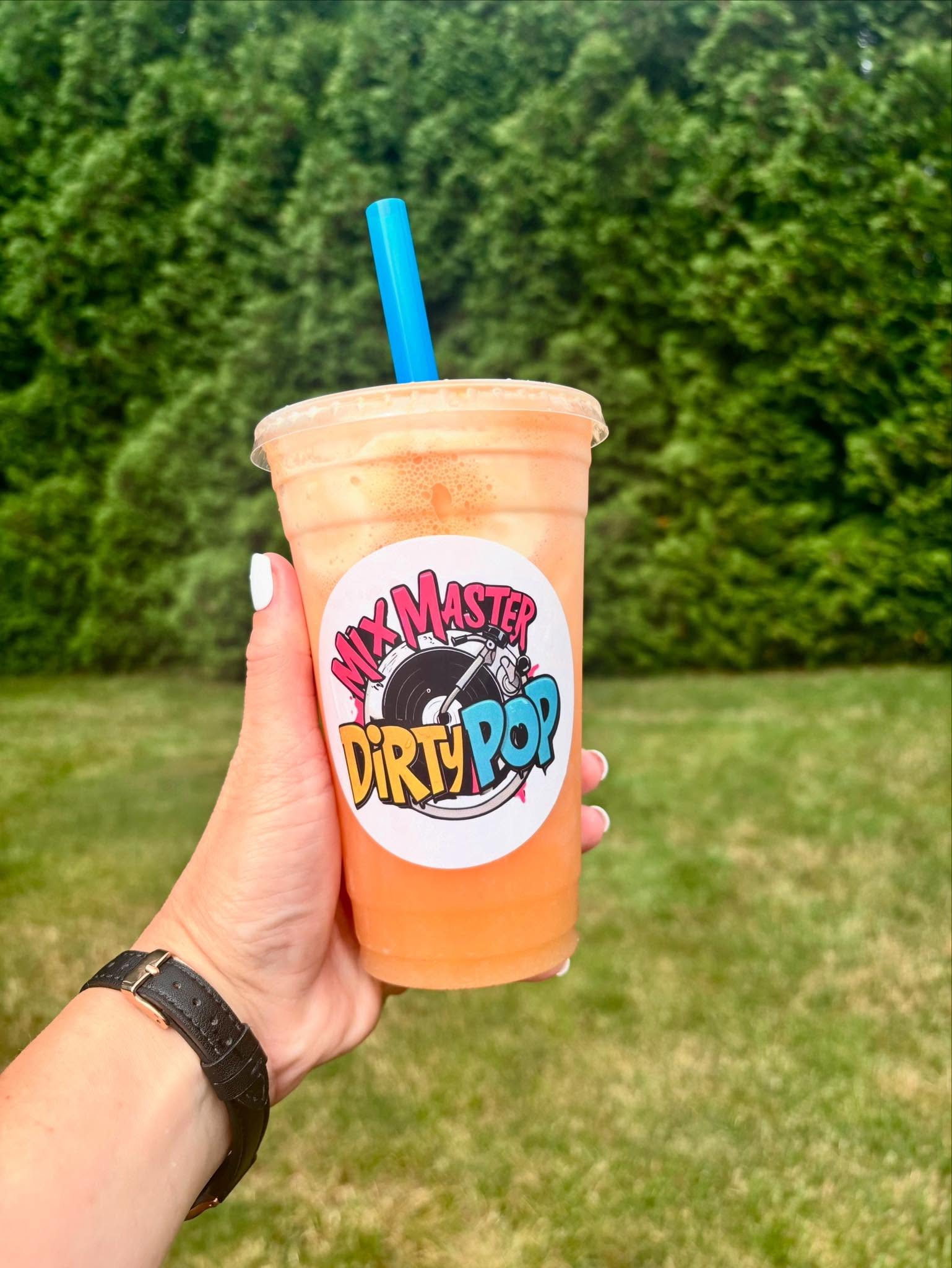 Hand holding a peach-colored smoothie with a blue straw outdoors, green trees in background.