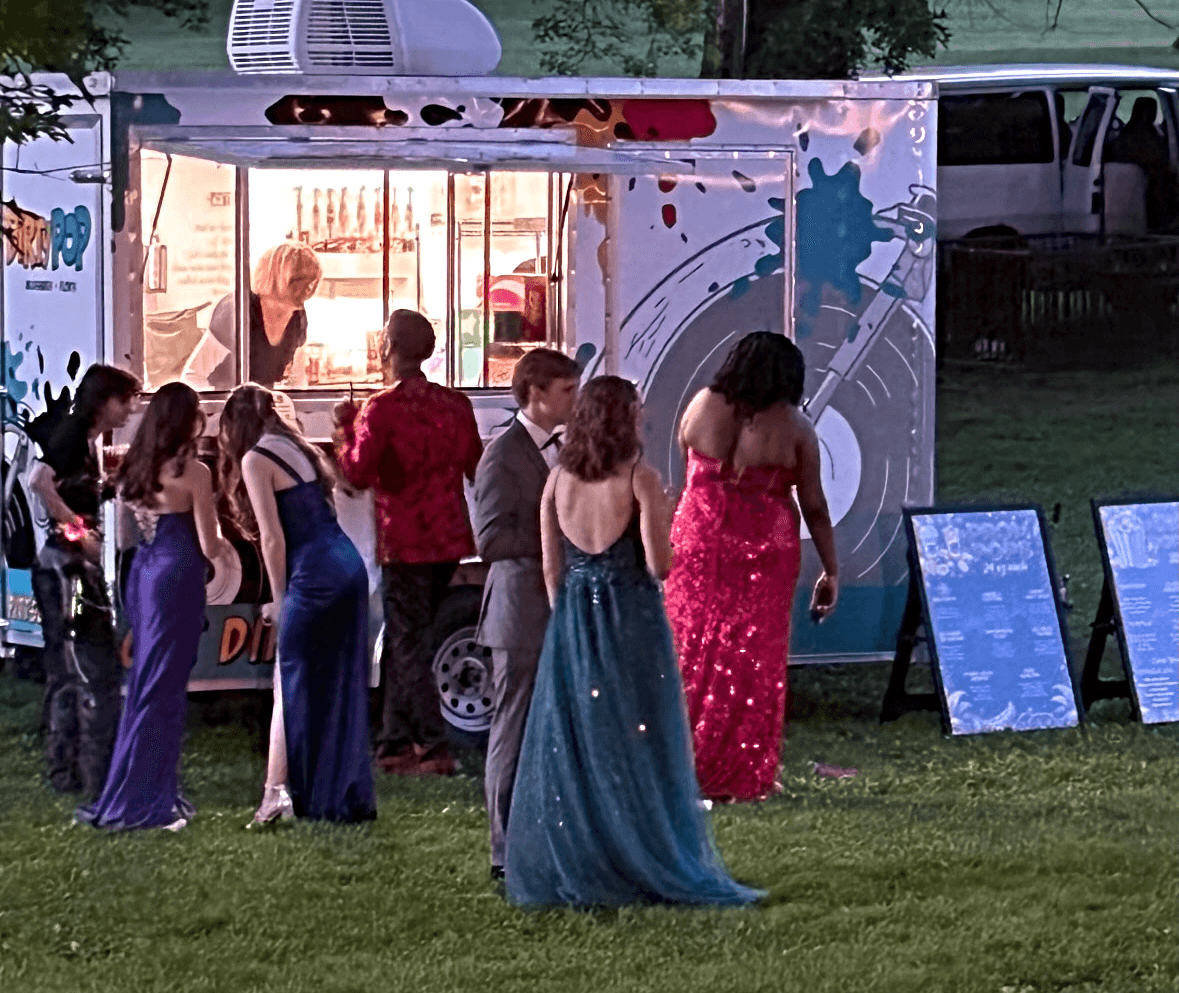 People in formal dresses line up outside an ice cream truck at dusk, with a grassy area and trees in background.