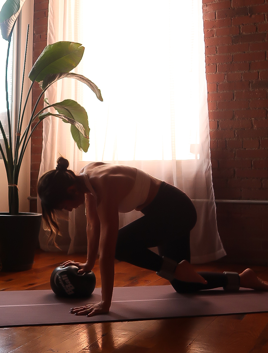 Person in a yoga pose on a mat near a large plant and window with curtains, brick wall in background.