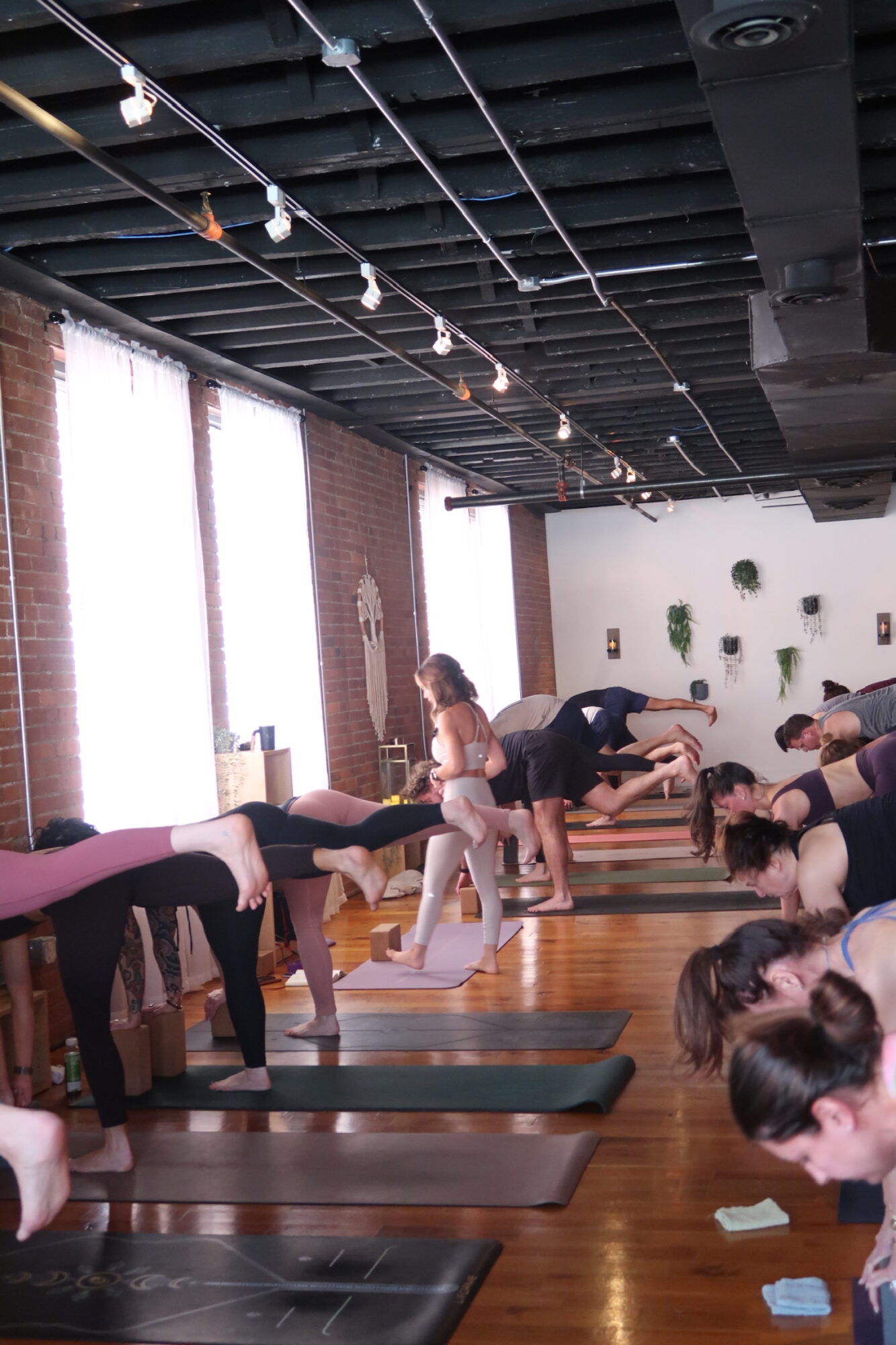 People participating in a yoga class in a studio with large windows, brick walls, and wooden floors.