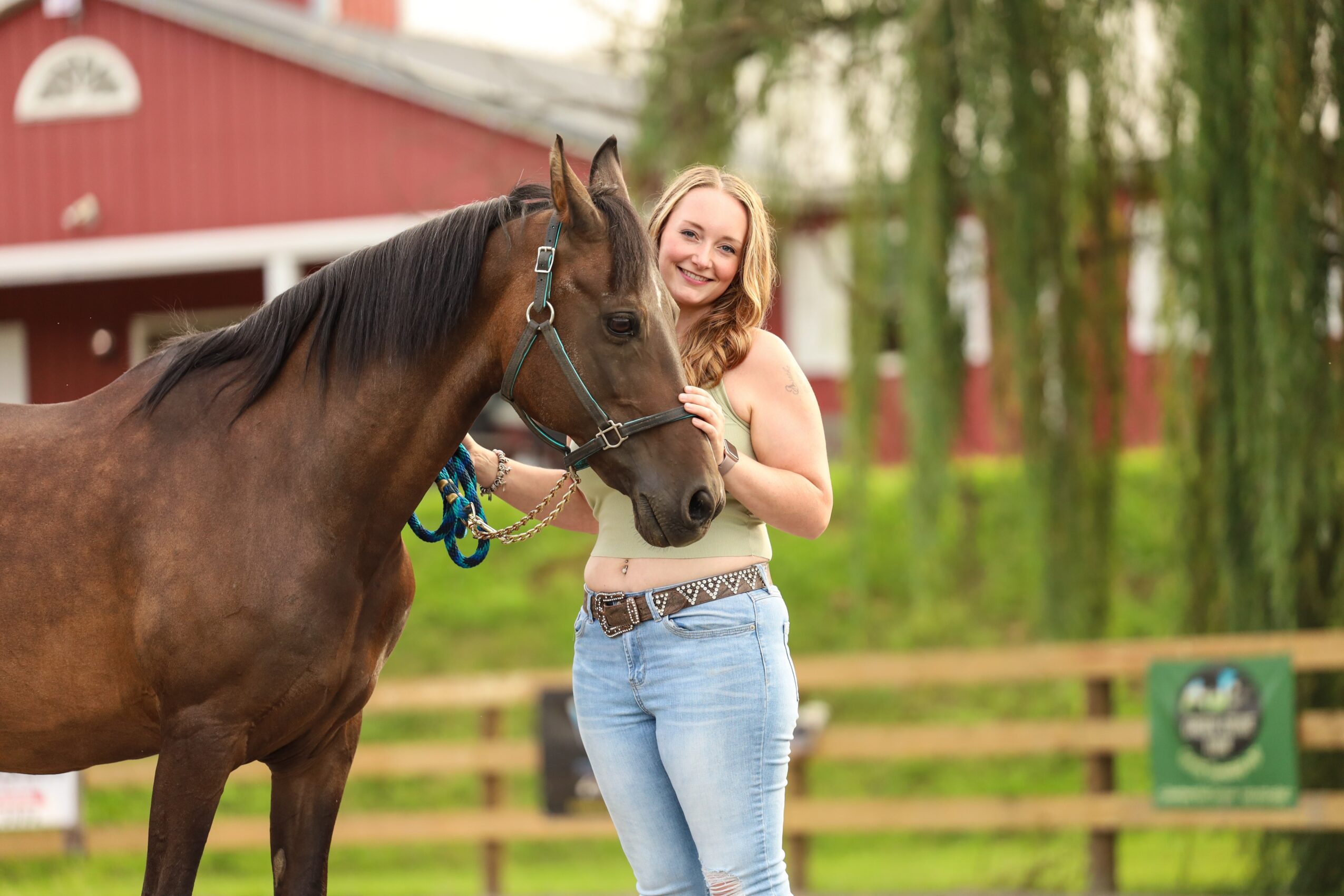 Young woman smiling next to a brown horse outdoors, with a red barn and green trees in the background.