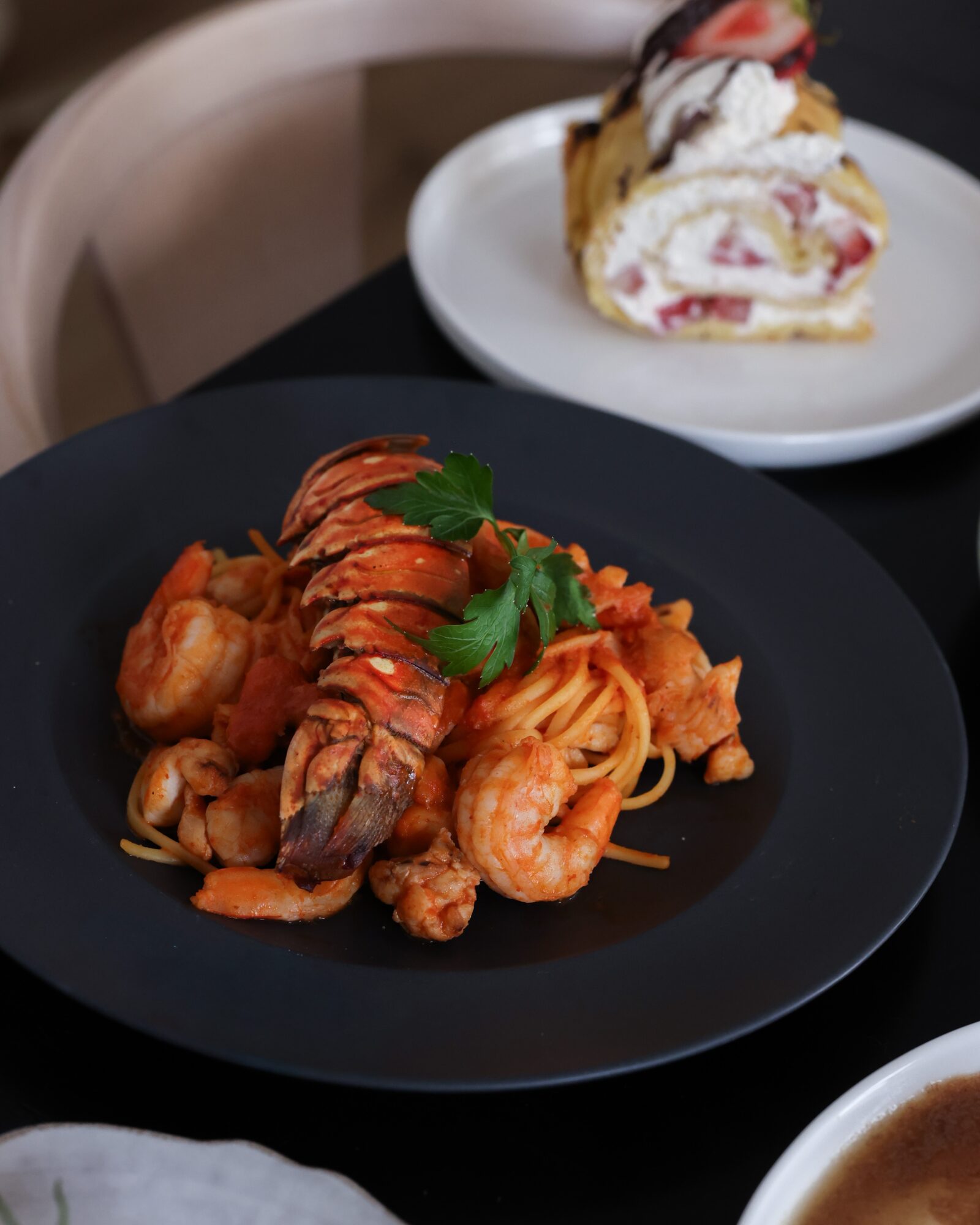 Plate with cooked shrimp, lobster tail, and pasta garnished with parsley, on a black plate.