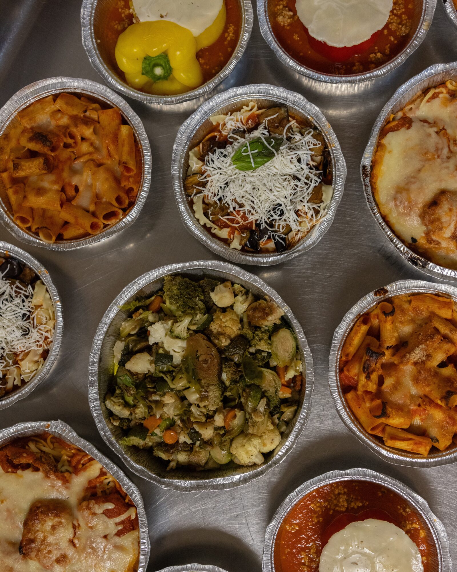 Assorted baked dishes in small aluminum containers, including pasta, vegetables, and cheese, arranged on a tray.