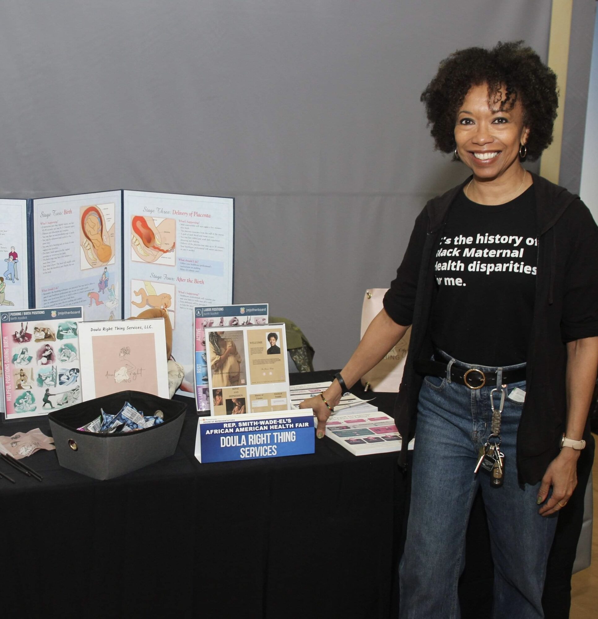 Smiling woman with curly hair standing next to a table with informational materials and posters, wearing a black T-shirt and jeans.