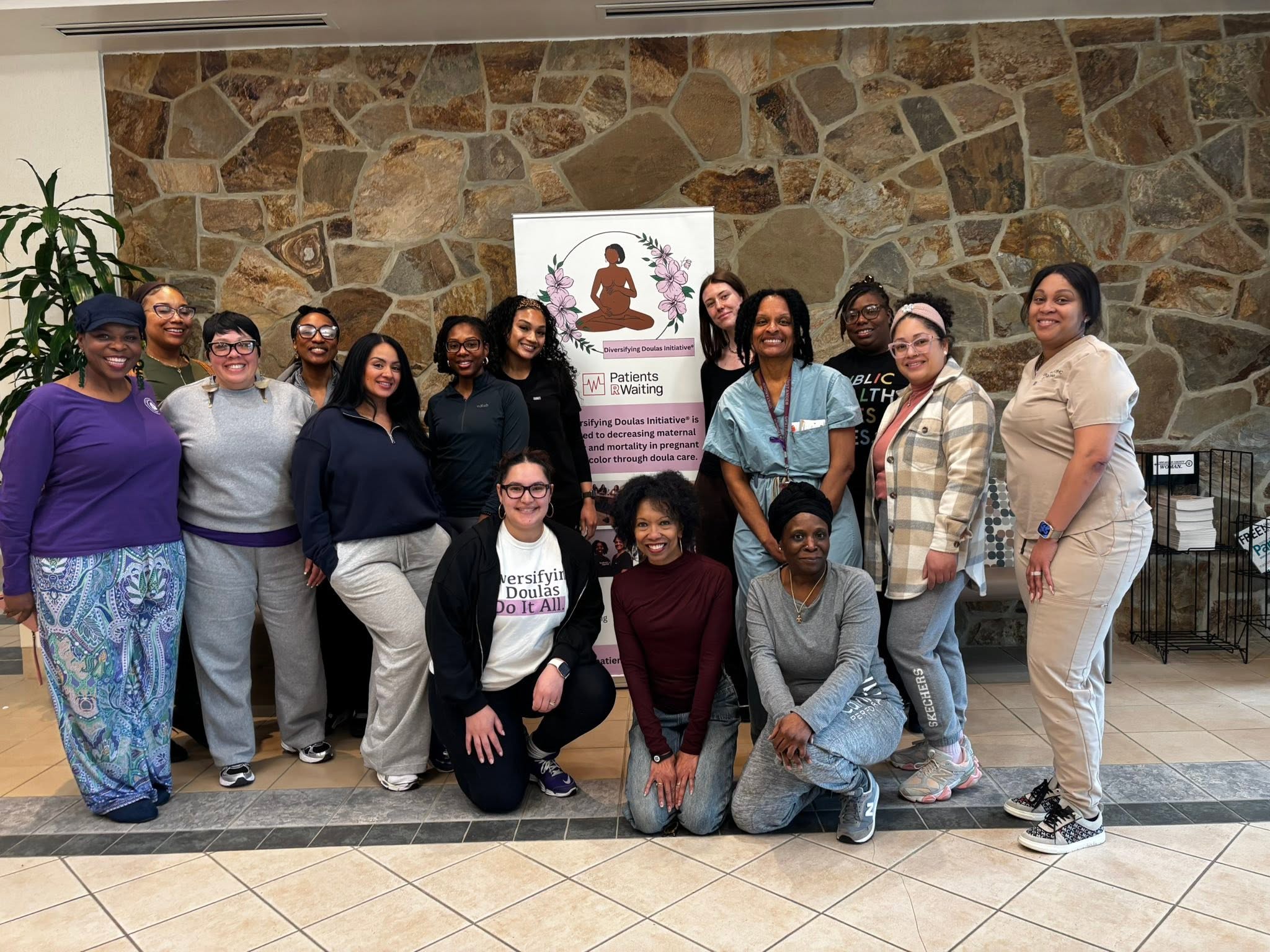 Group of 14 women standing indoors in front of a stone wall and a poster, smiling, with some kneeling in front.