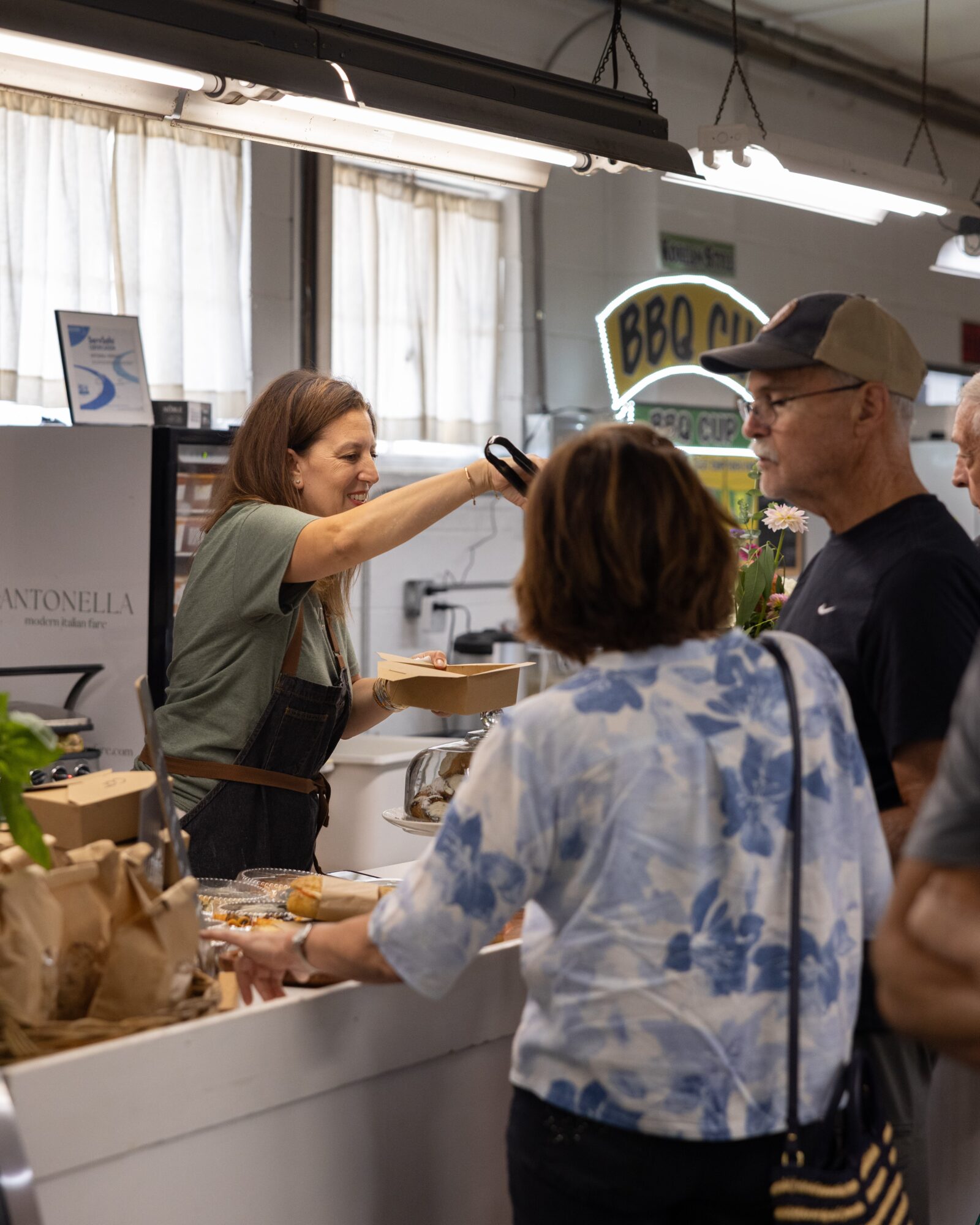 A woman at a food counter hands a receipt to a customer, with people waiting nearby and a BBQ sign in the background.