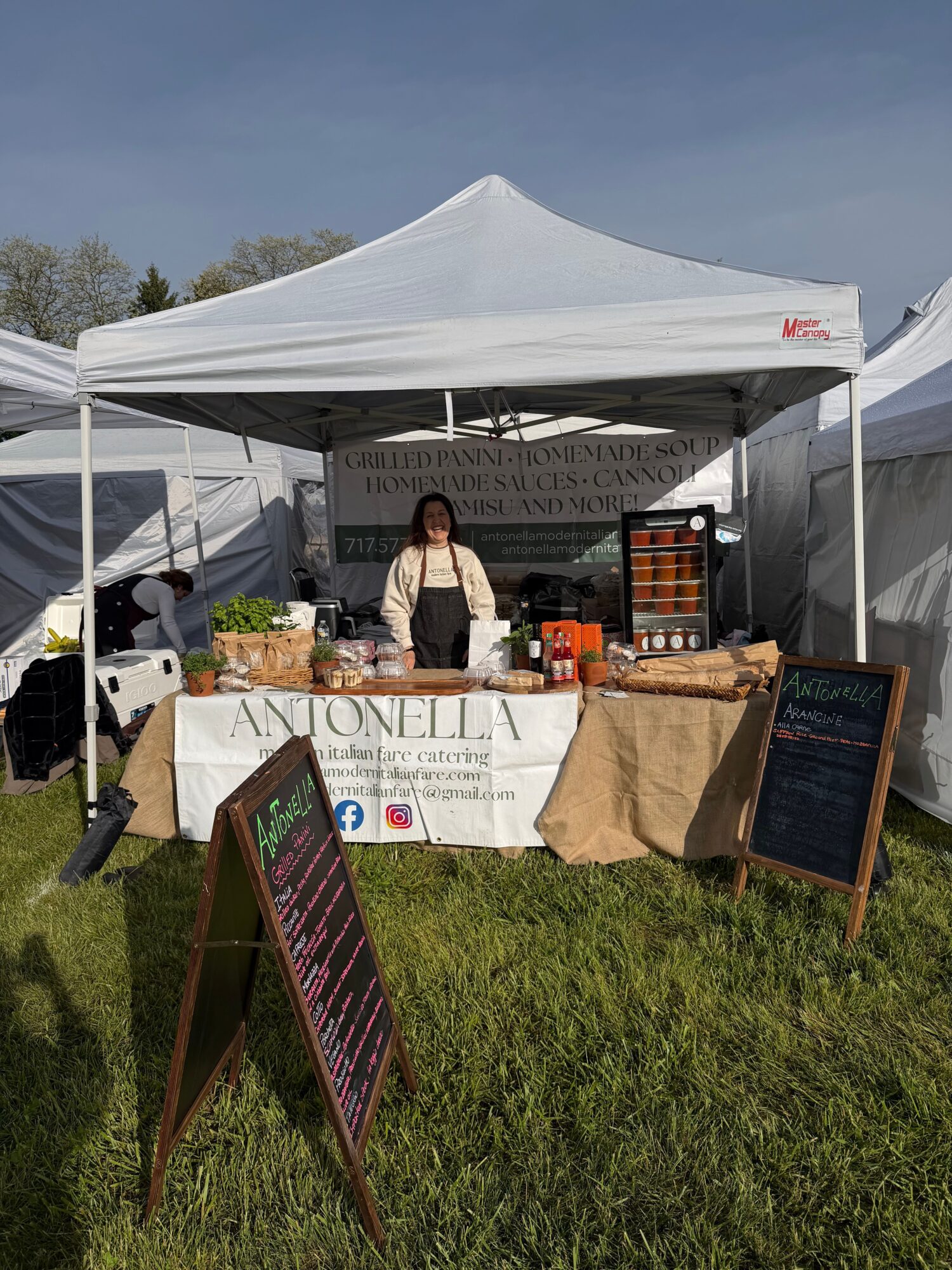 Woman standing behind a table under a white canopy at an outdoor event, with signs and items on display.