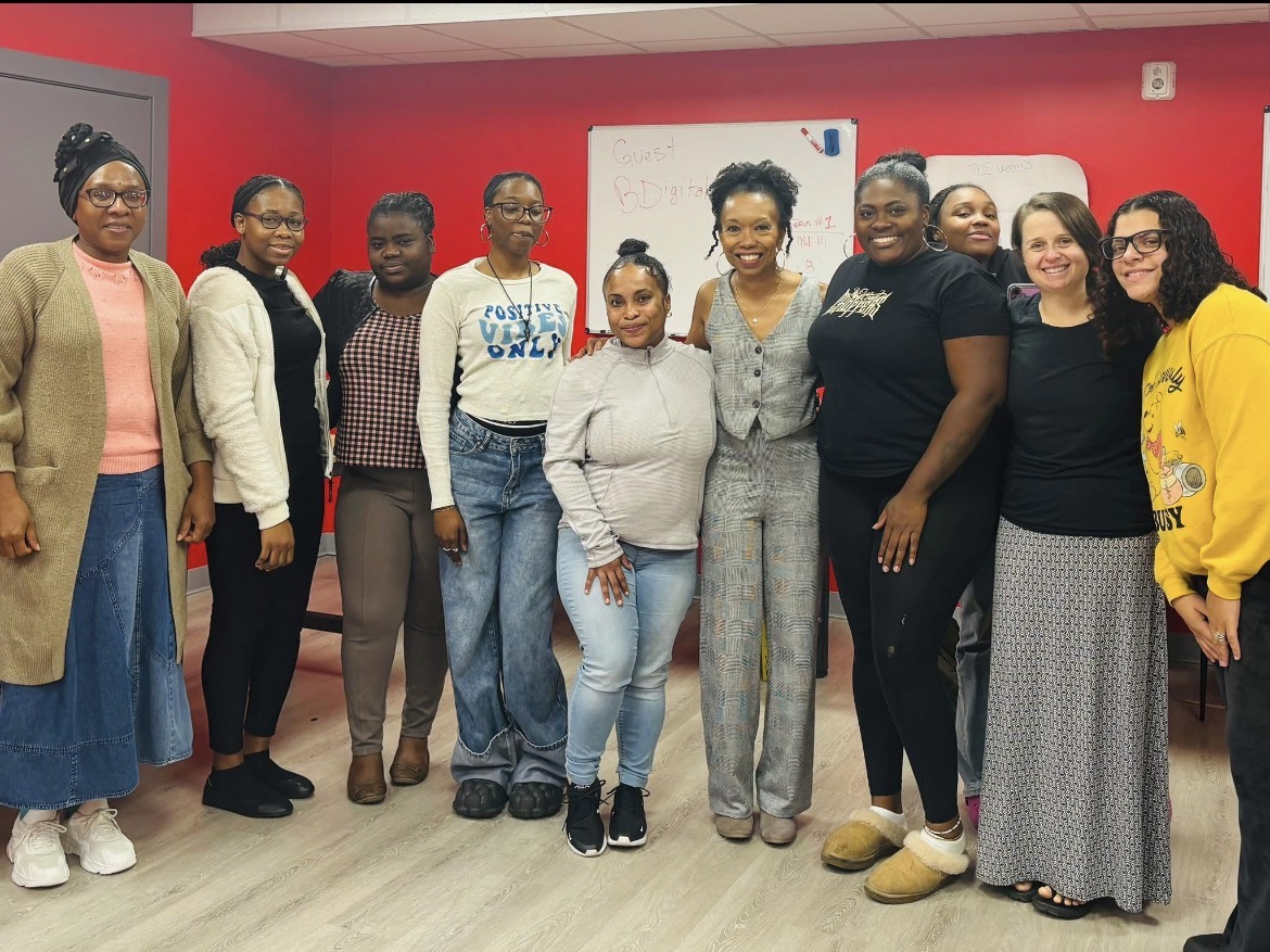 Group of eleven women standing indoors against a red wall, smiling, with a whiteboard behind them.