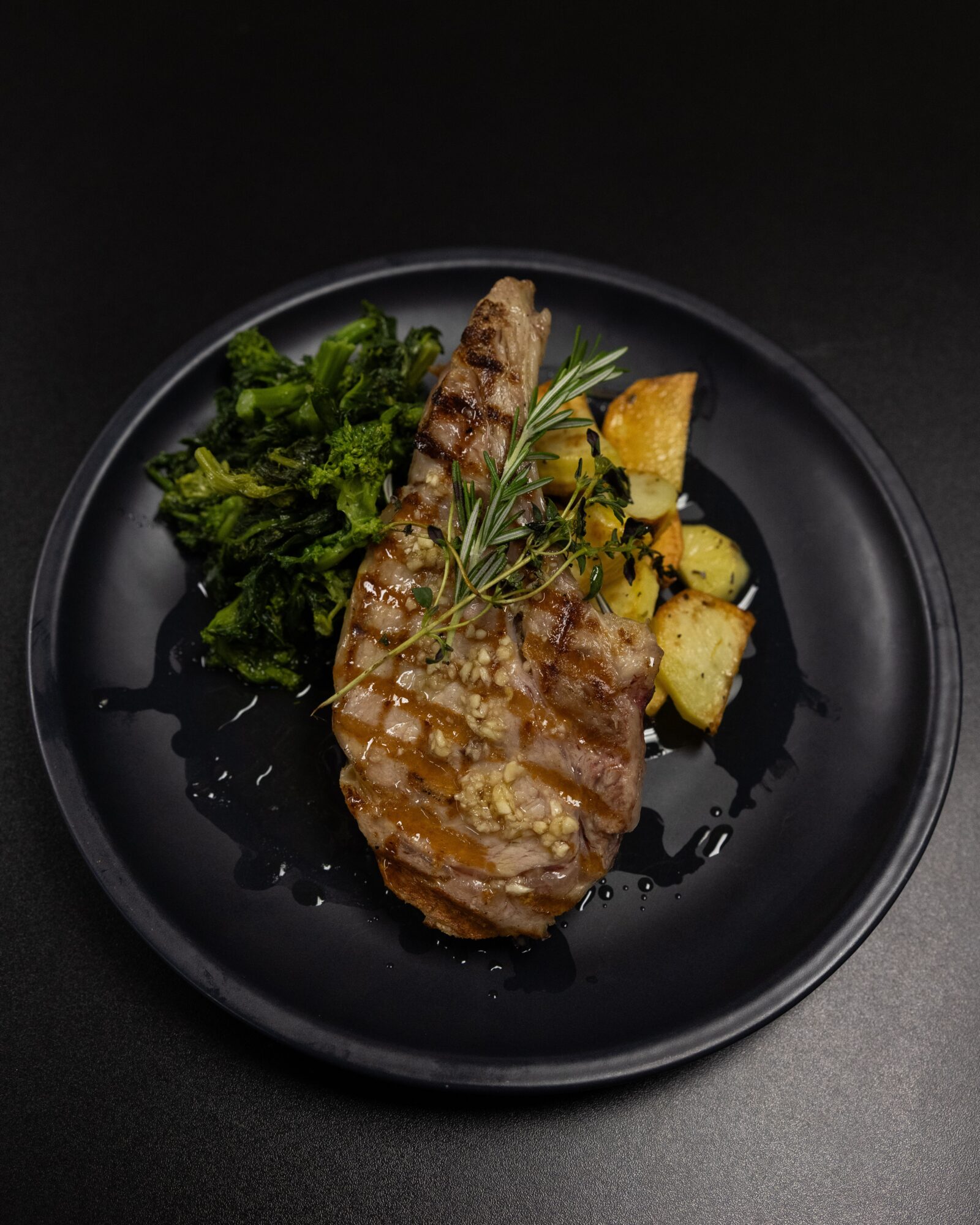Grilled meat with herbs, roasted potatoes, and greens on a black plate against a dark background.