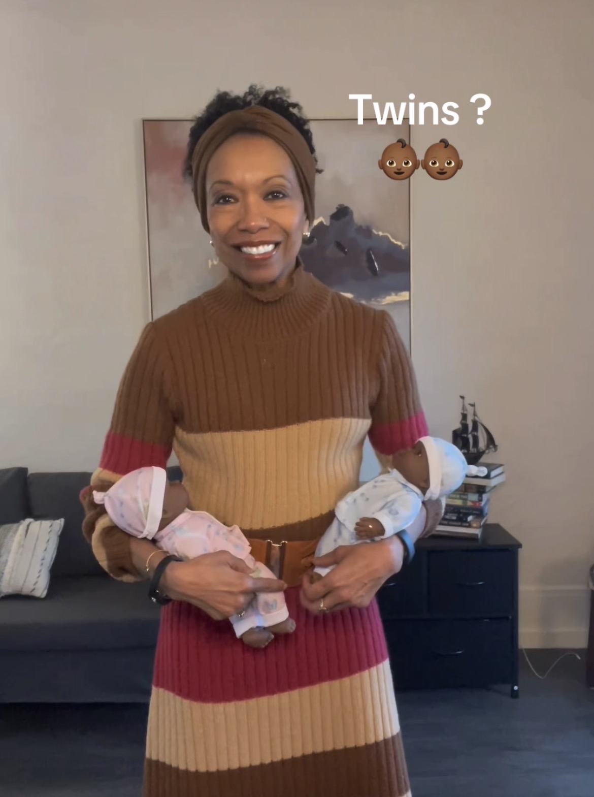 Woman smiling holding two babies, one in each arm, with a painting and furniture in background.