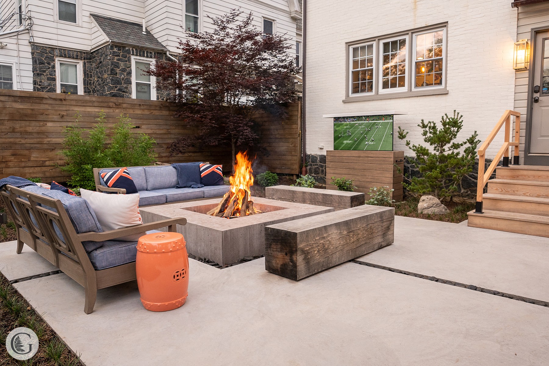 Backyard patio with fire pit, seating, plants, and stairs leading to a house, during evening with lit window.